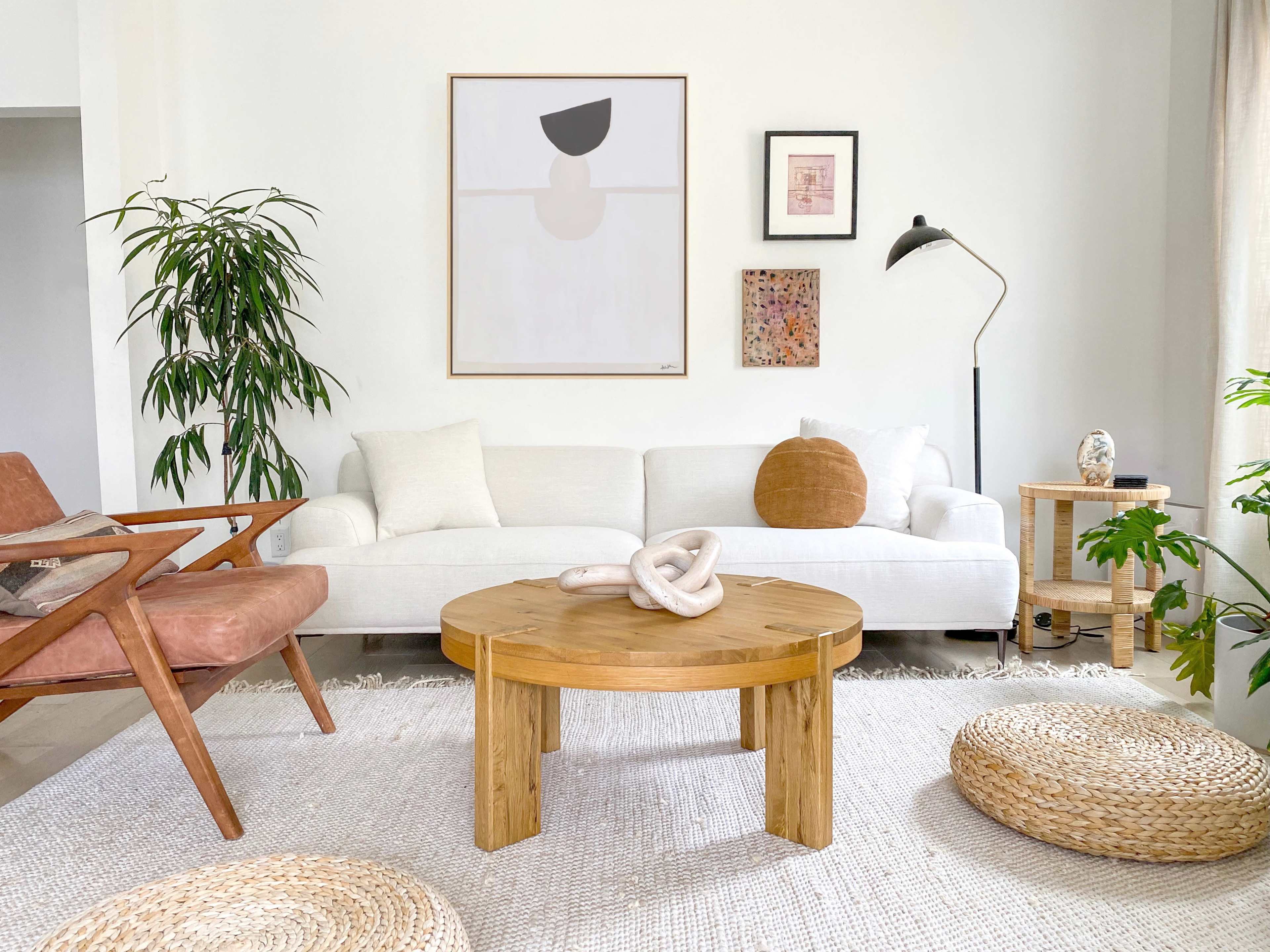 A bright living room features a white couch, a round wooden coffee table, a large rug, and various decorative elements, including plants and artwork on the walls.