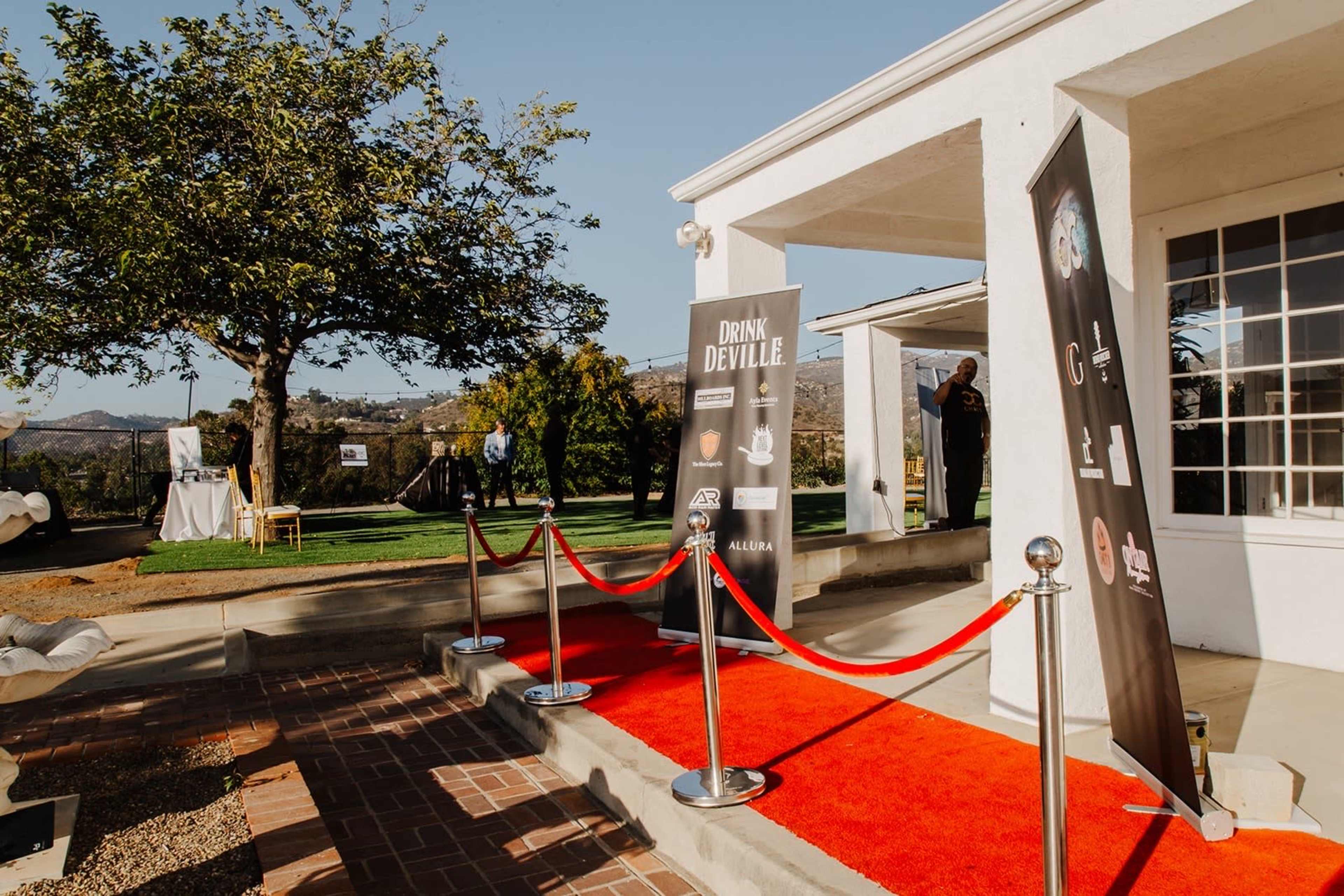 A red carpet leads to a white building with banners and a tree in the background on a sunny day.