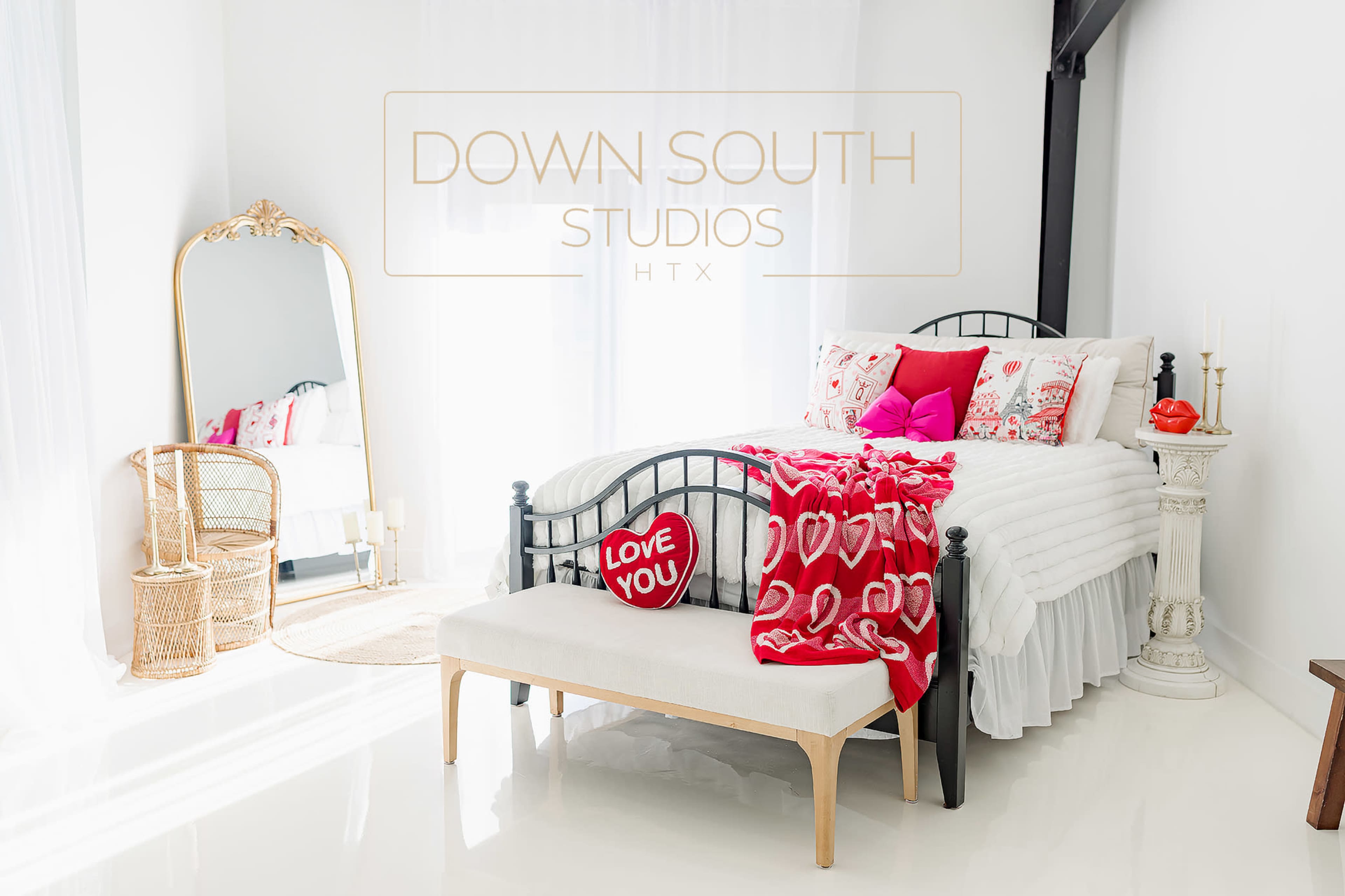 The image depicts a bright and airy bedroom featuring a black metal bed adorned with decorative pillows and a red blanket, a large mirror, and a white bench.