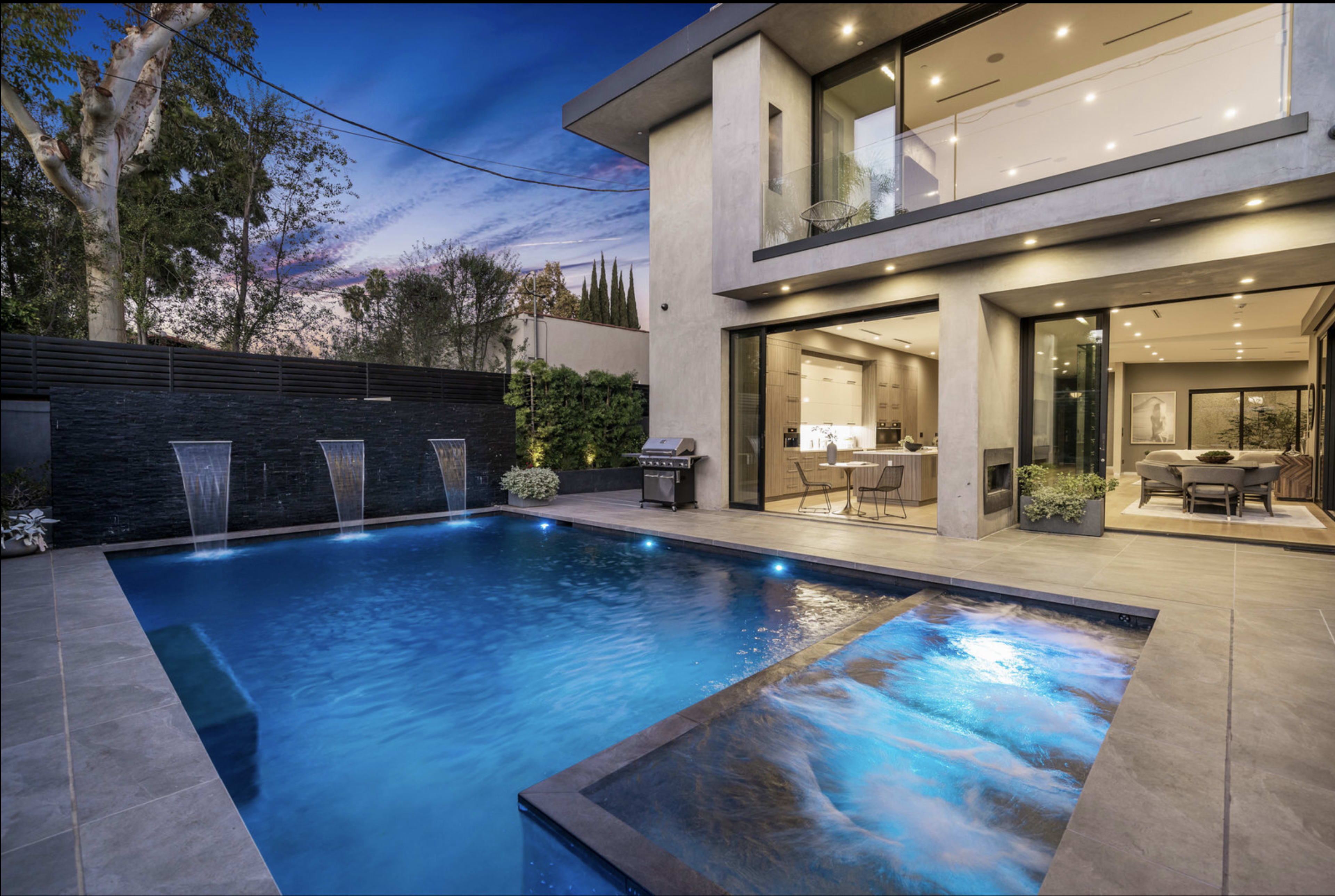 A modern home features a pool with water fountains and a terrace surrounded by greenery, illuminated by evening lights.