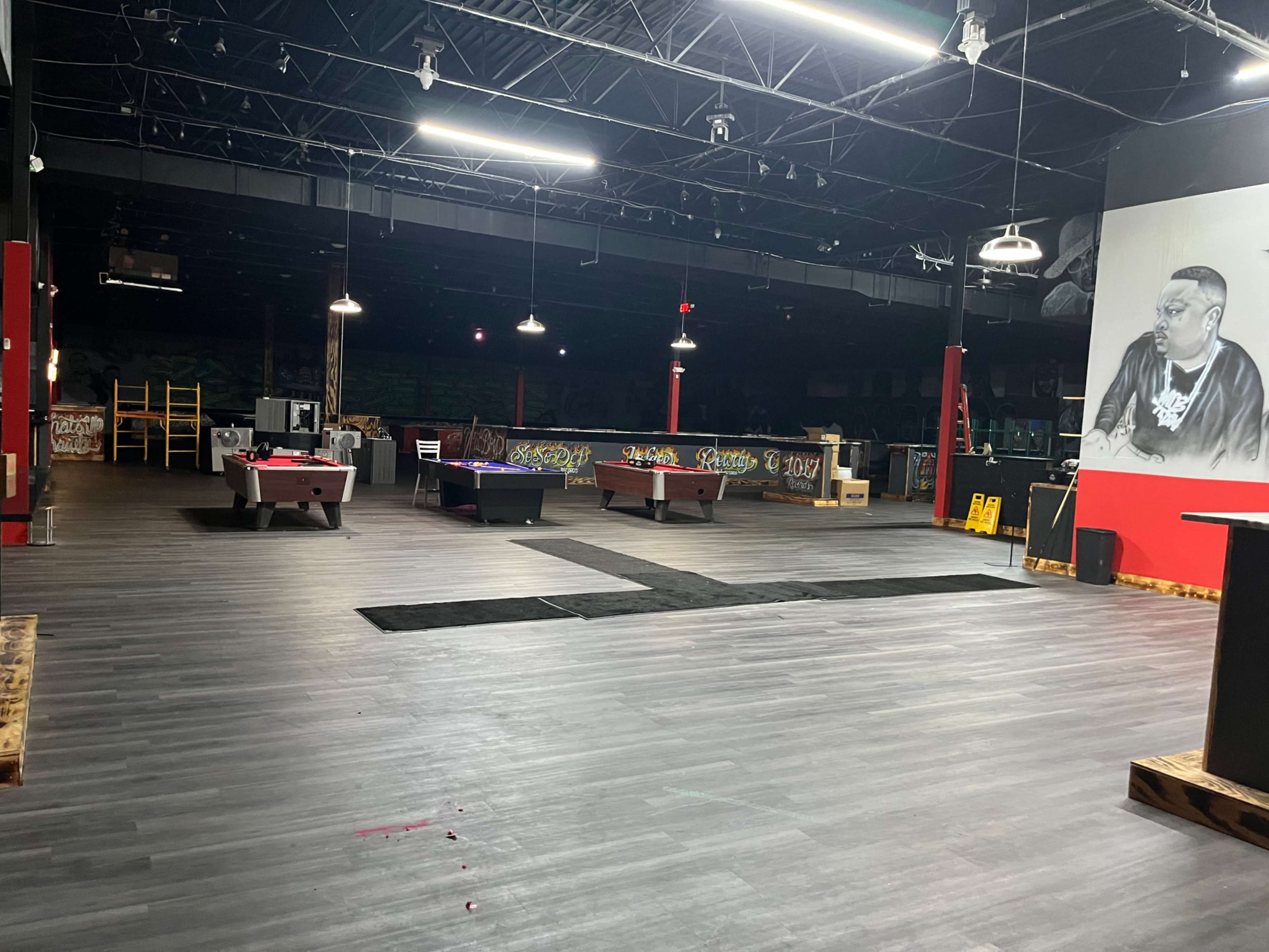 The image shows a large, empty indoor space with several pool tables, industrial lighting, and graffiti on the walls.