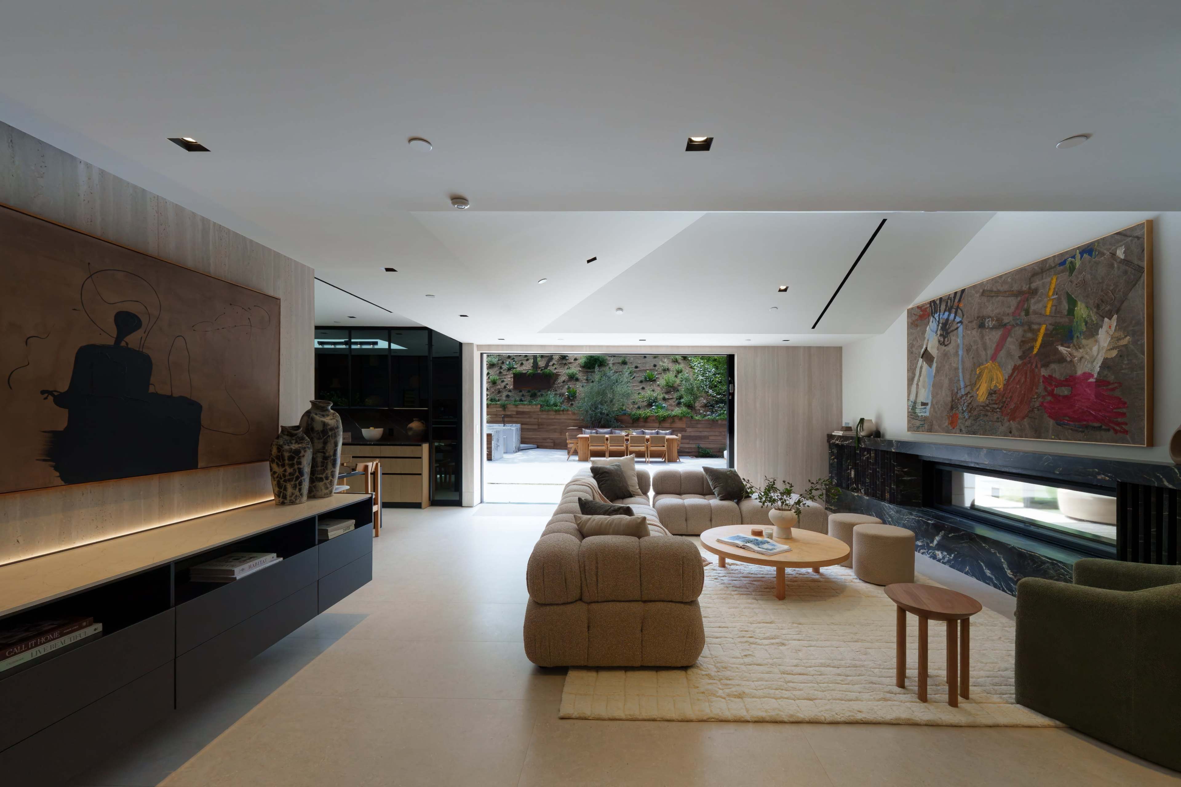 The image shows a modern living room with a large sectional sofa, two abstract paintings on the walls, and a glass door leading to an outdoor patio area.