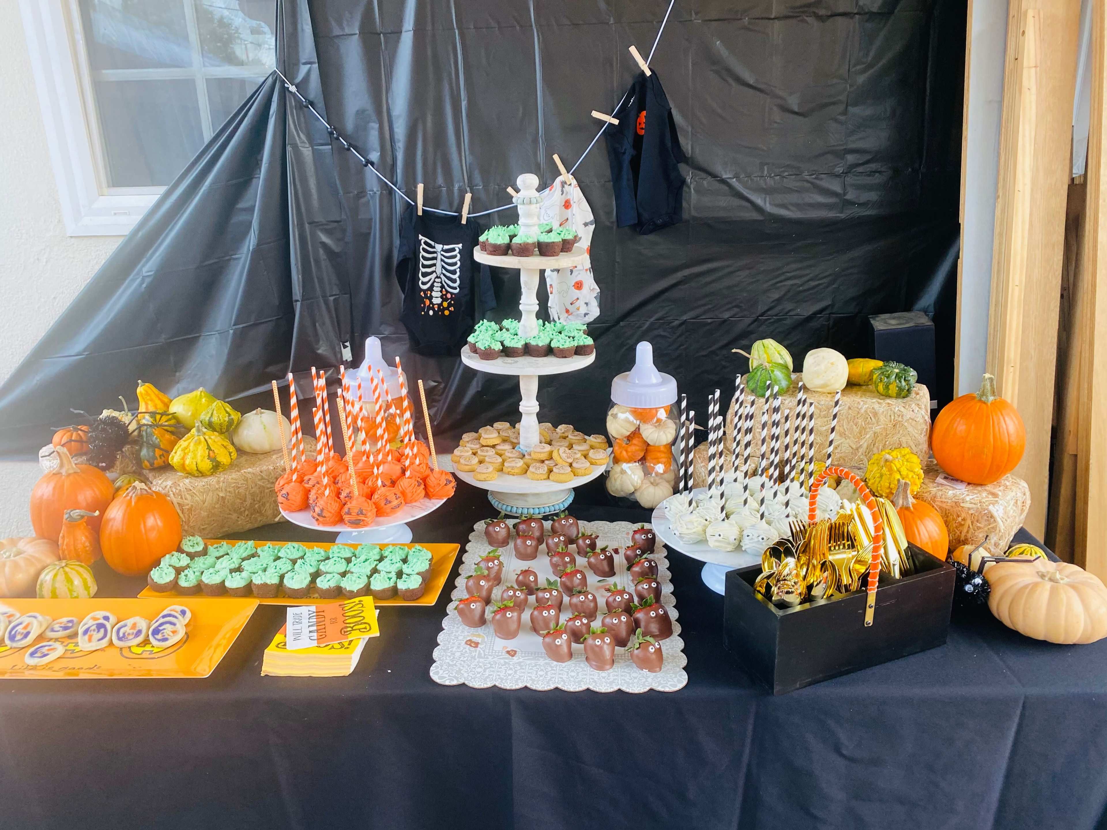 A table is decorated with various Halloween-themed treats, including cupcakes, candy pumpkins, and decorative gourds, set against a black backdrop.