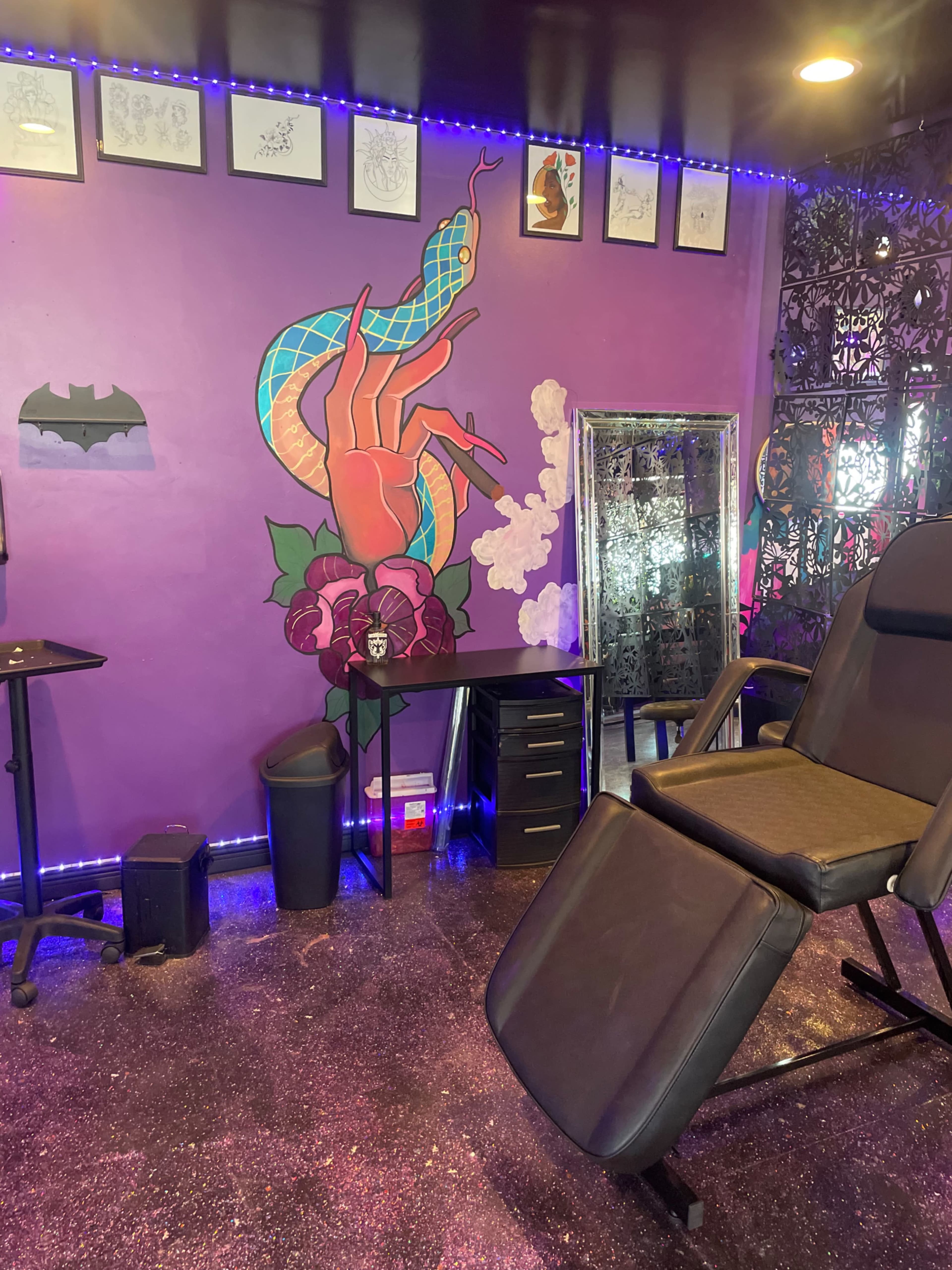 The image shows a tattoo studio with a purple wall featuring a serpent and floral mural, a tattoo chair, a desk, and framed sketches on display.