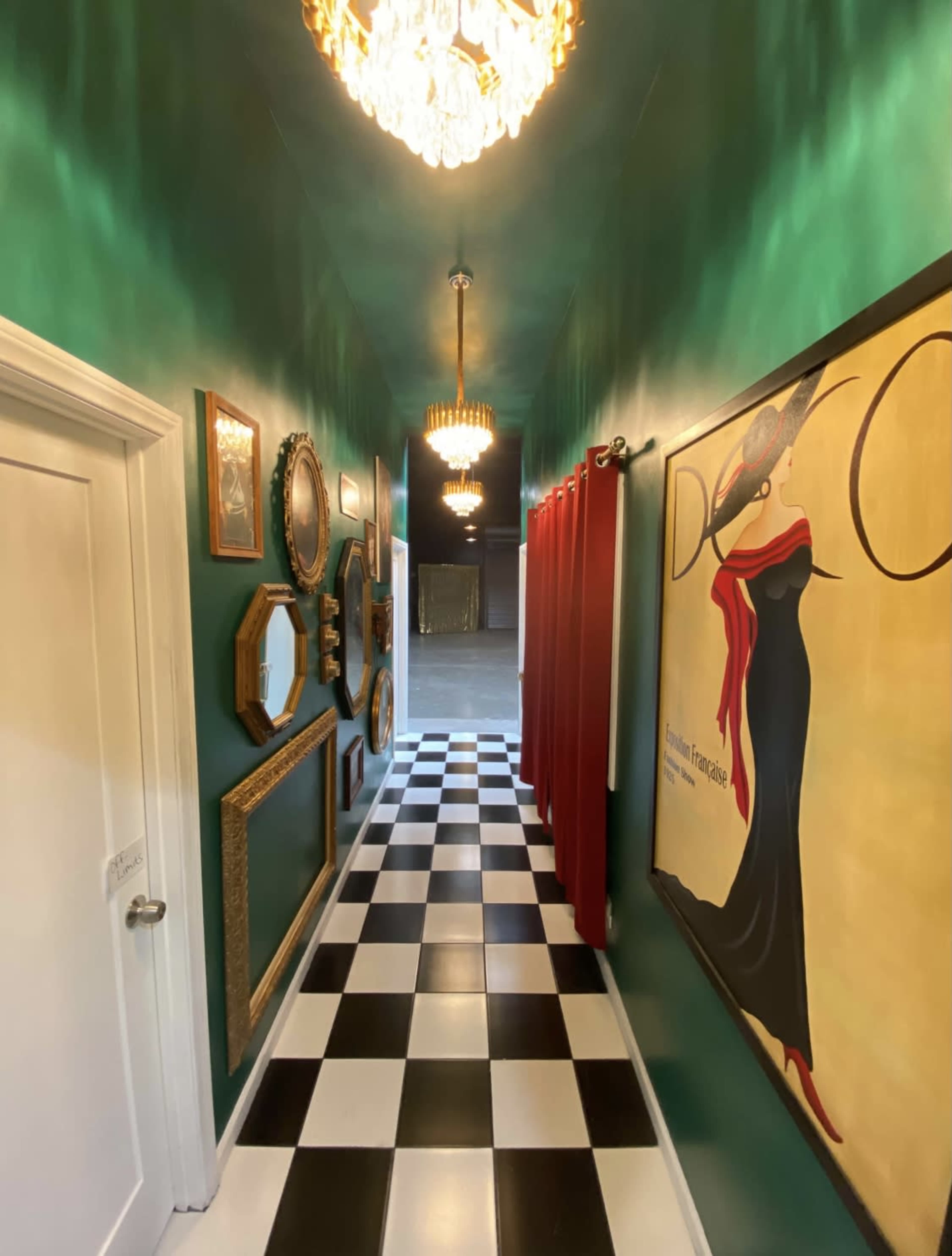 The image shows a narrow hallway with green walls, black and white checkered flooring, framed mirrors, and a large decorative painting, illuminated by pendant lights.