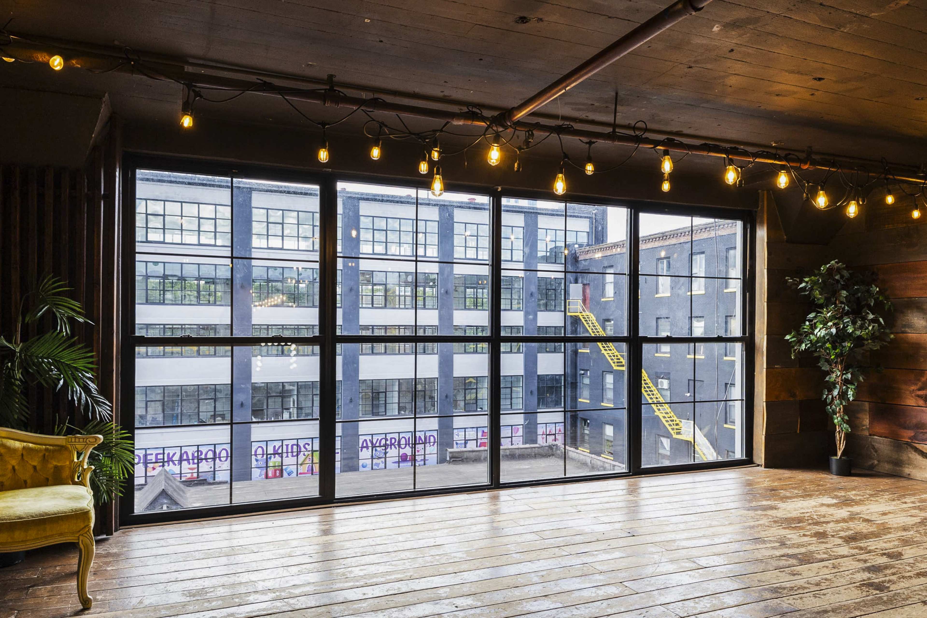 A spacious interior room features large windows overlooking a neighboring building with yellow fire escape stairs.