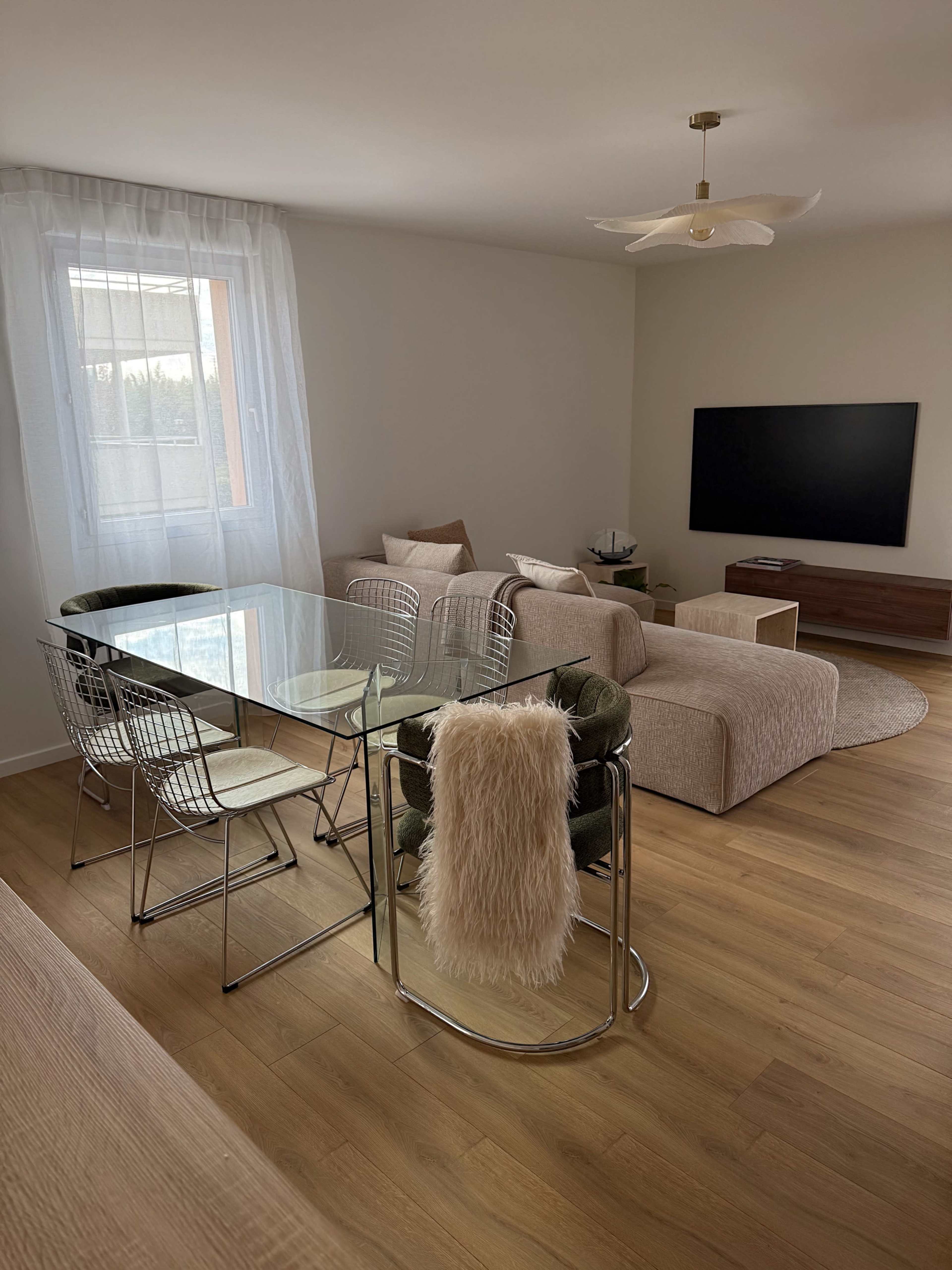 The image shows a modern living room with a glass dining table surrounded by wire chairs, a beige sofa, a wall-mounted television, and a large window with sheer curtains.