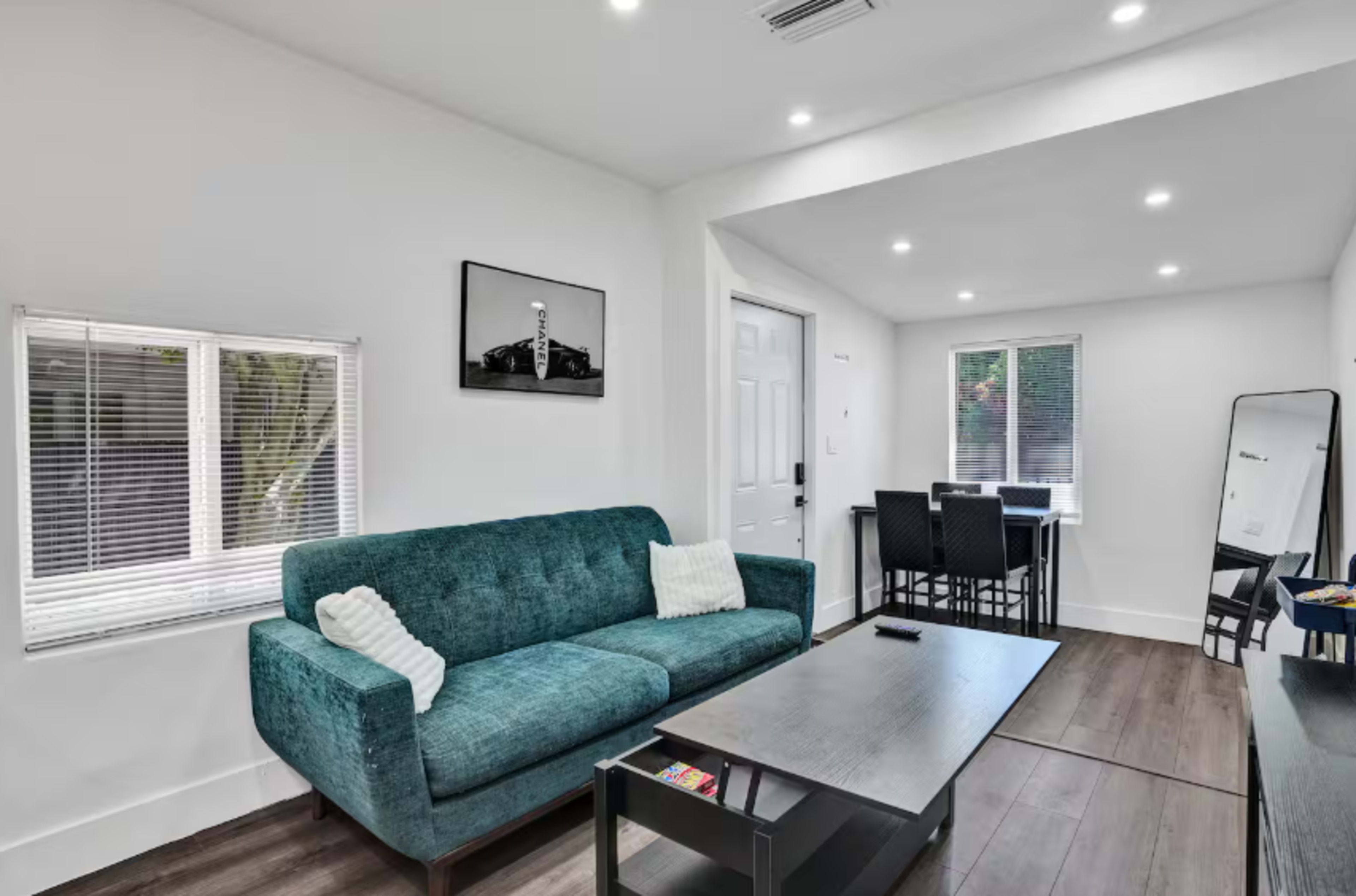 The image shows a modern living area with a teal sofa, a coffee table, and a dining table with six black chairs, all set against white walls and bright lighting.