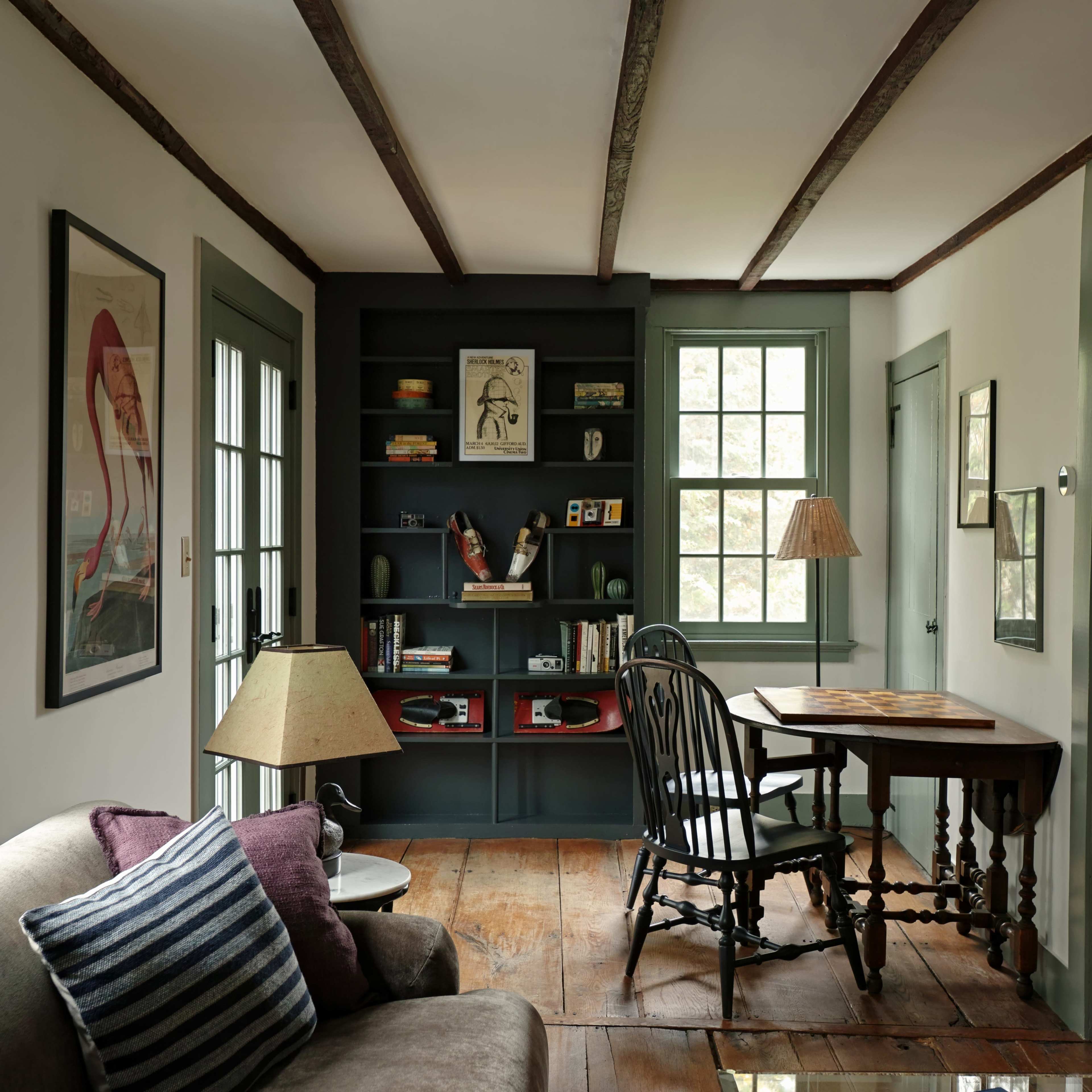 The image shows a cozy room with a wooden floor, a small table and chair, a bookshelf filled with various items, and large windows allowing natural light to enter.