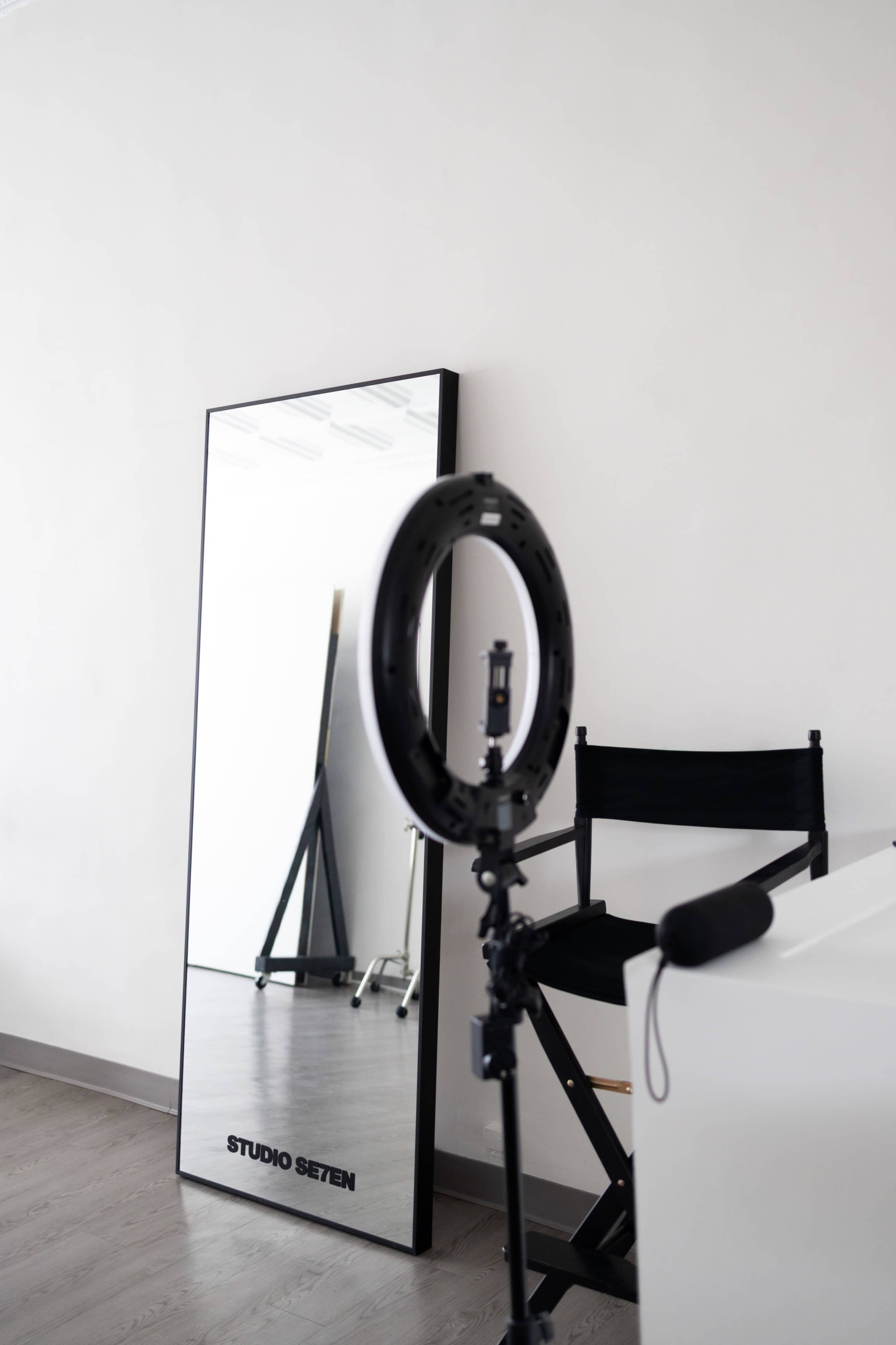 The image shows a minimalist studio setup featuring a black director's chair, a ring light on a stand, and a large mirror against a white wall.