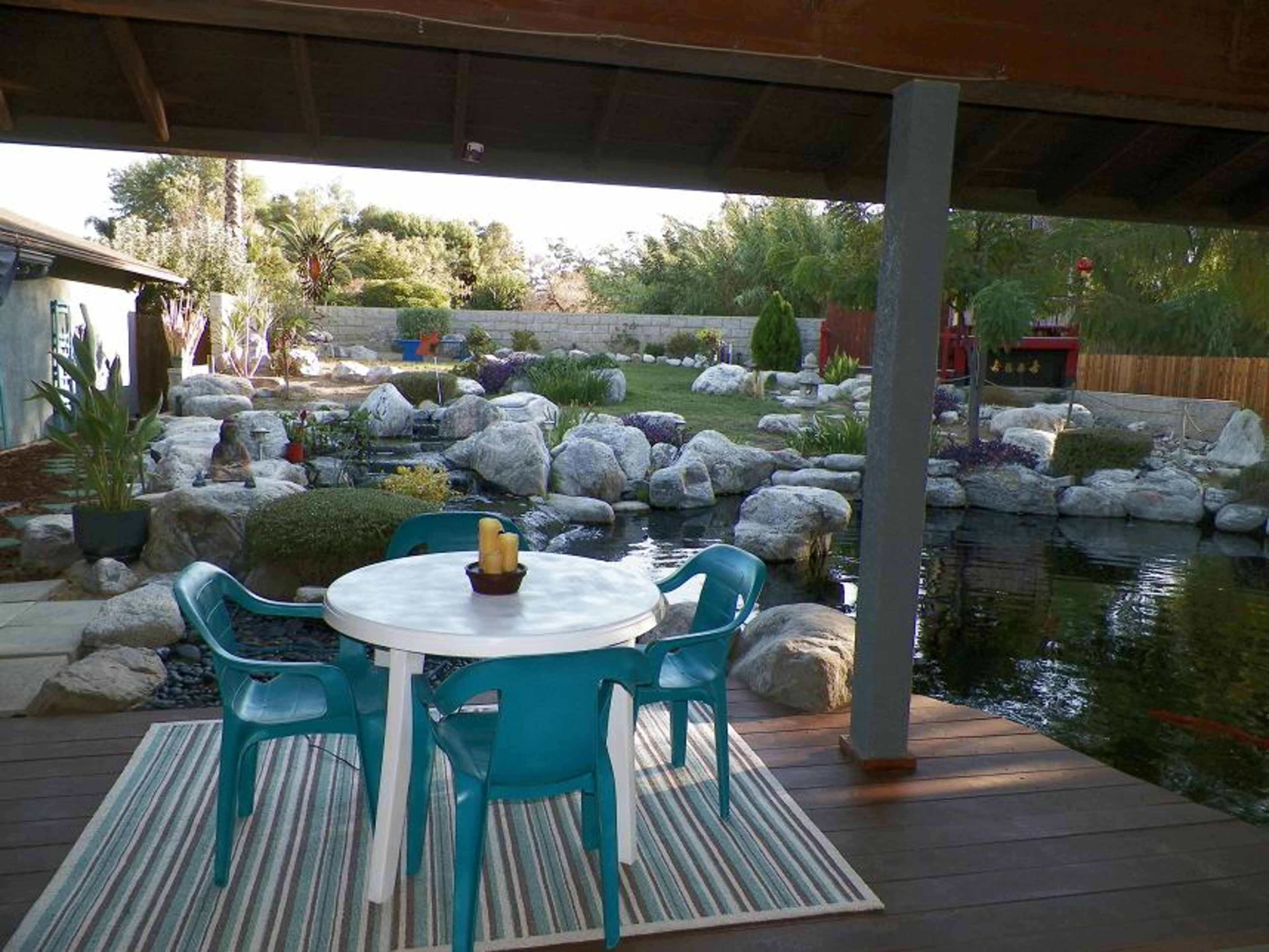 A dining table with blue chairs sits on a wooden patio overlooking a landscaped yard with a pond and rock features.