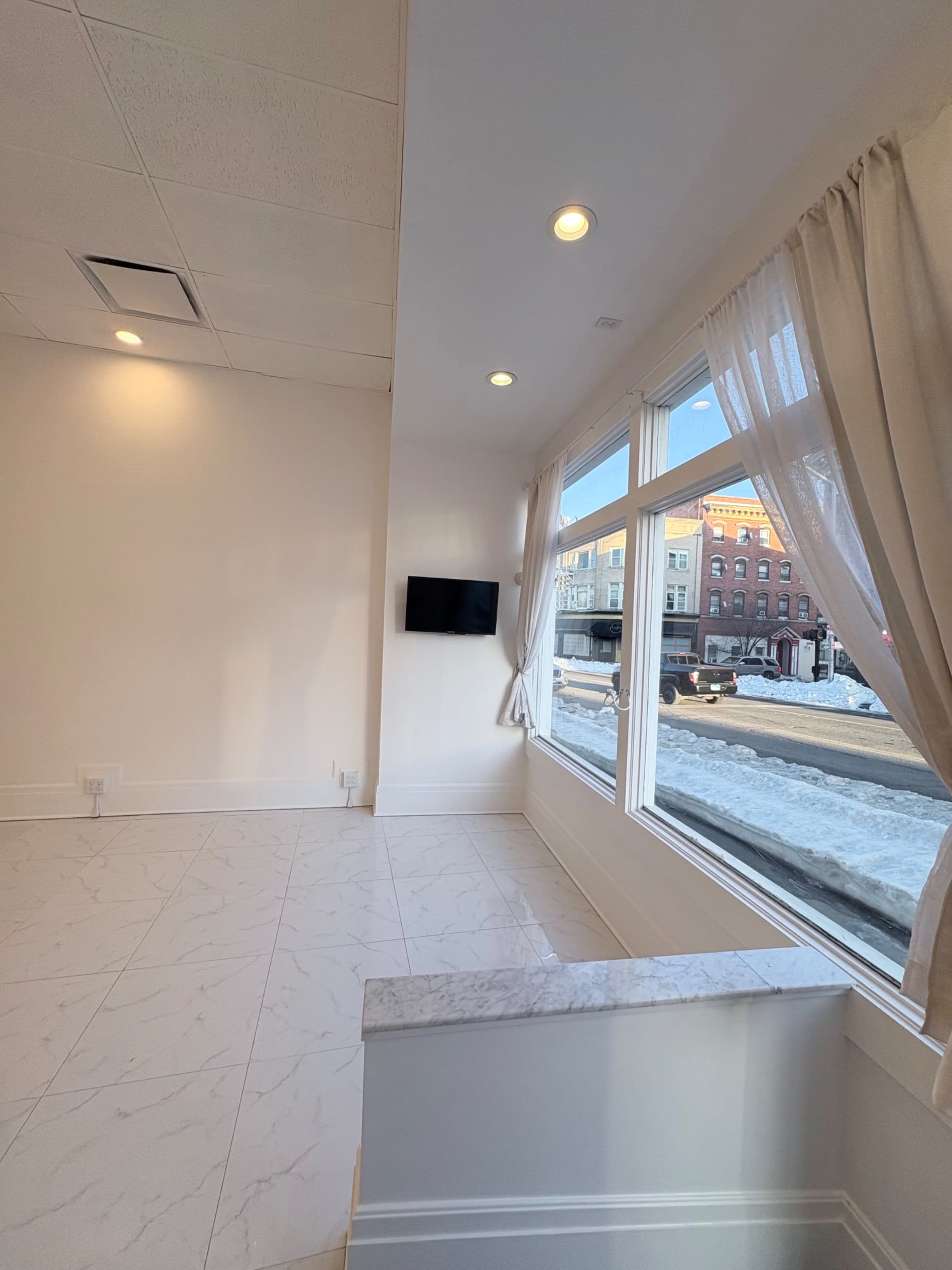 The image shows a bright, modern interior space with large windows, white walls, and a flat-screen TV mounted on one wall.