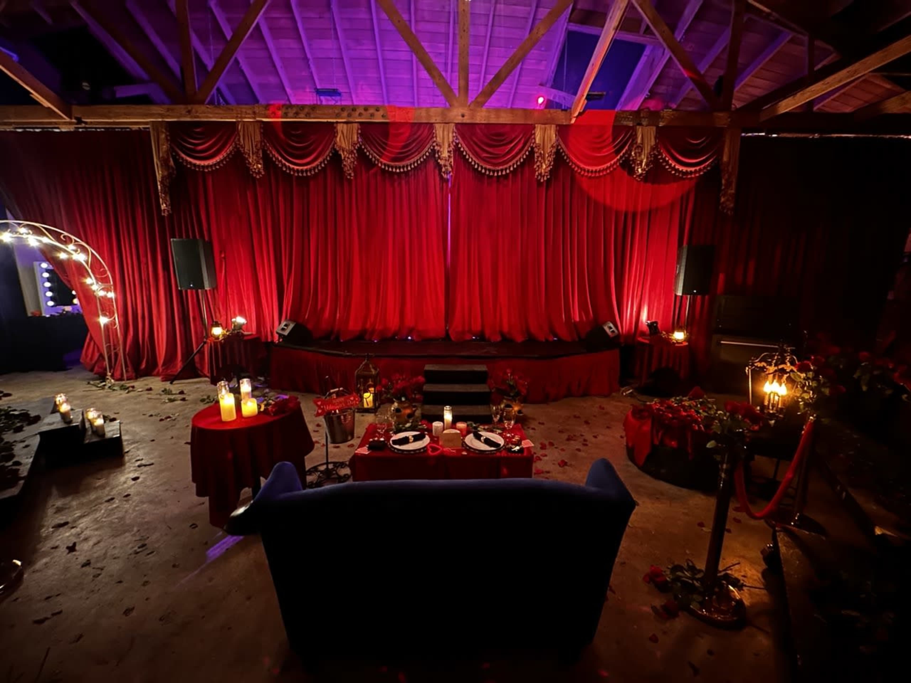 A dimly lit event space with red drapery, tables set with candles and dinnerware, and a stage in the background.