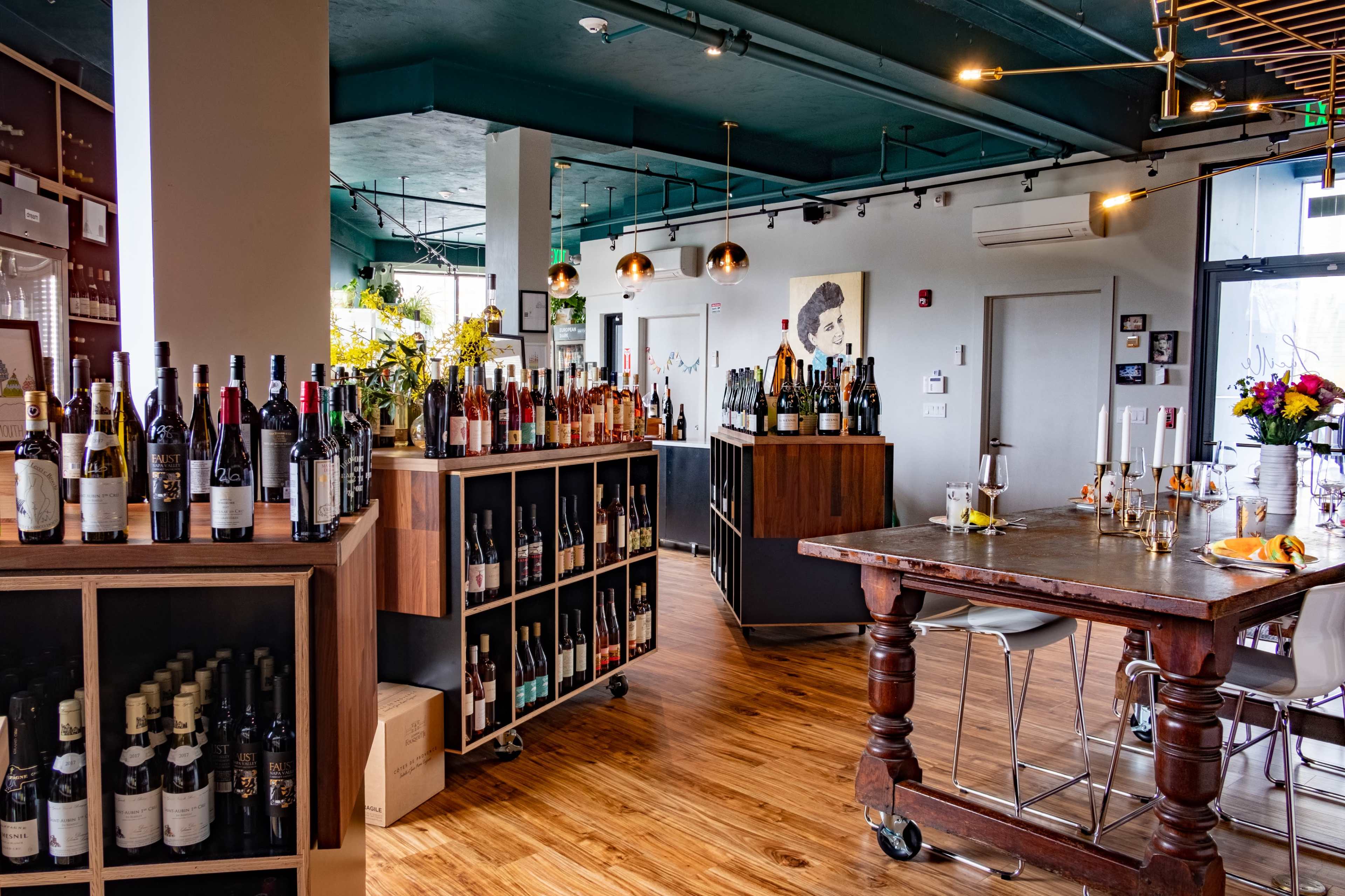 A modern wine bar featuring wooden shelves filled with various bottles of wine, a large wooden table in the center, and floral arrangements enhancing the ambiance.