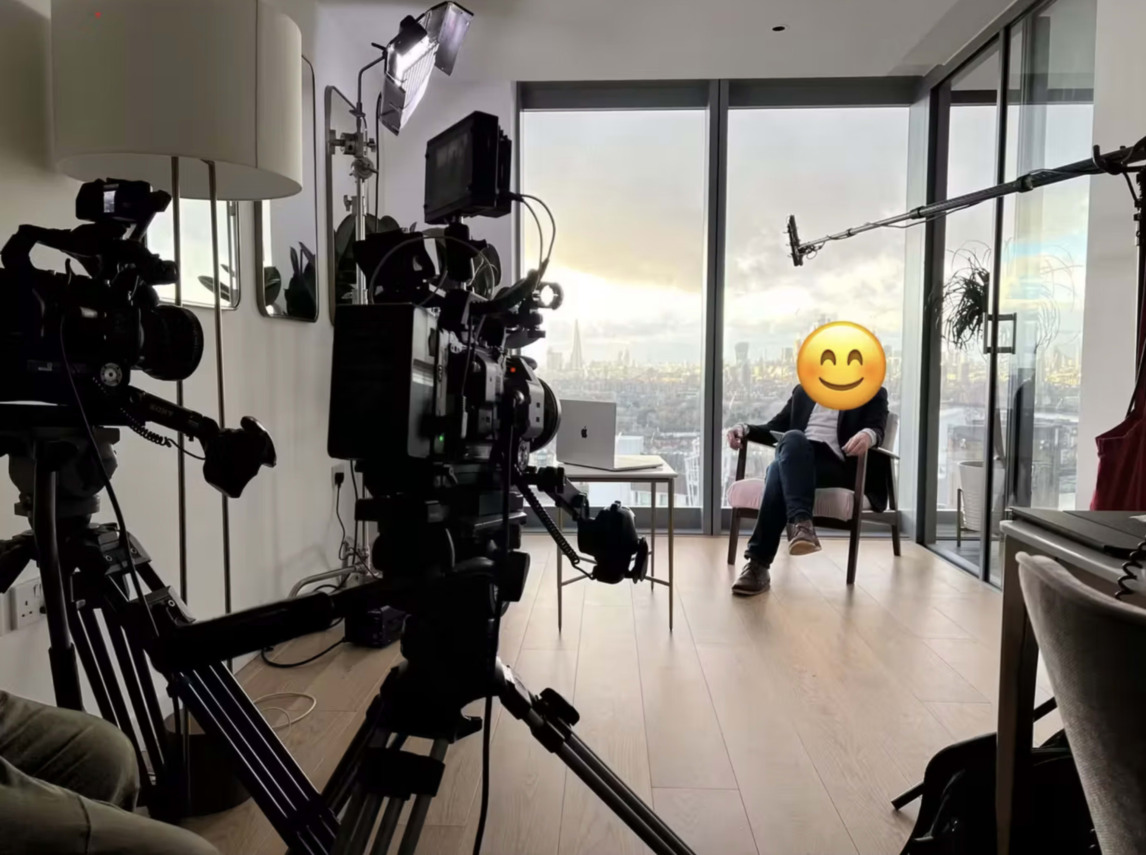 Two cameras are positioned in a modern room with large windows showcasing a city skyline, while a person sits in a chair with an emoji covering their face.