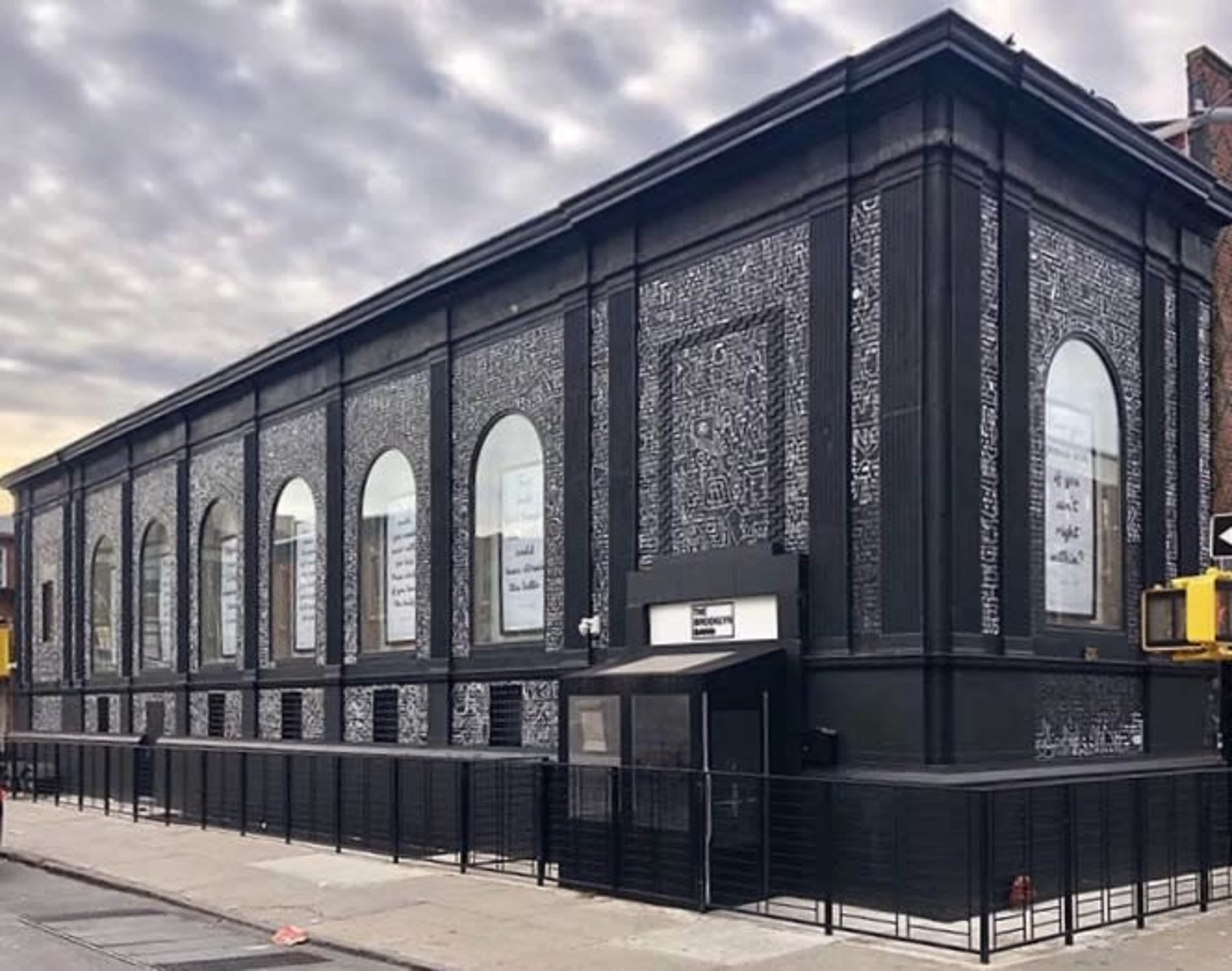 A large, black-painted building with multiple arched windows featuring white text and a fenced sidewalk in front.