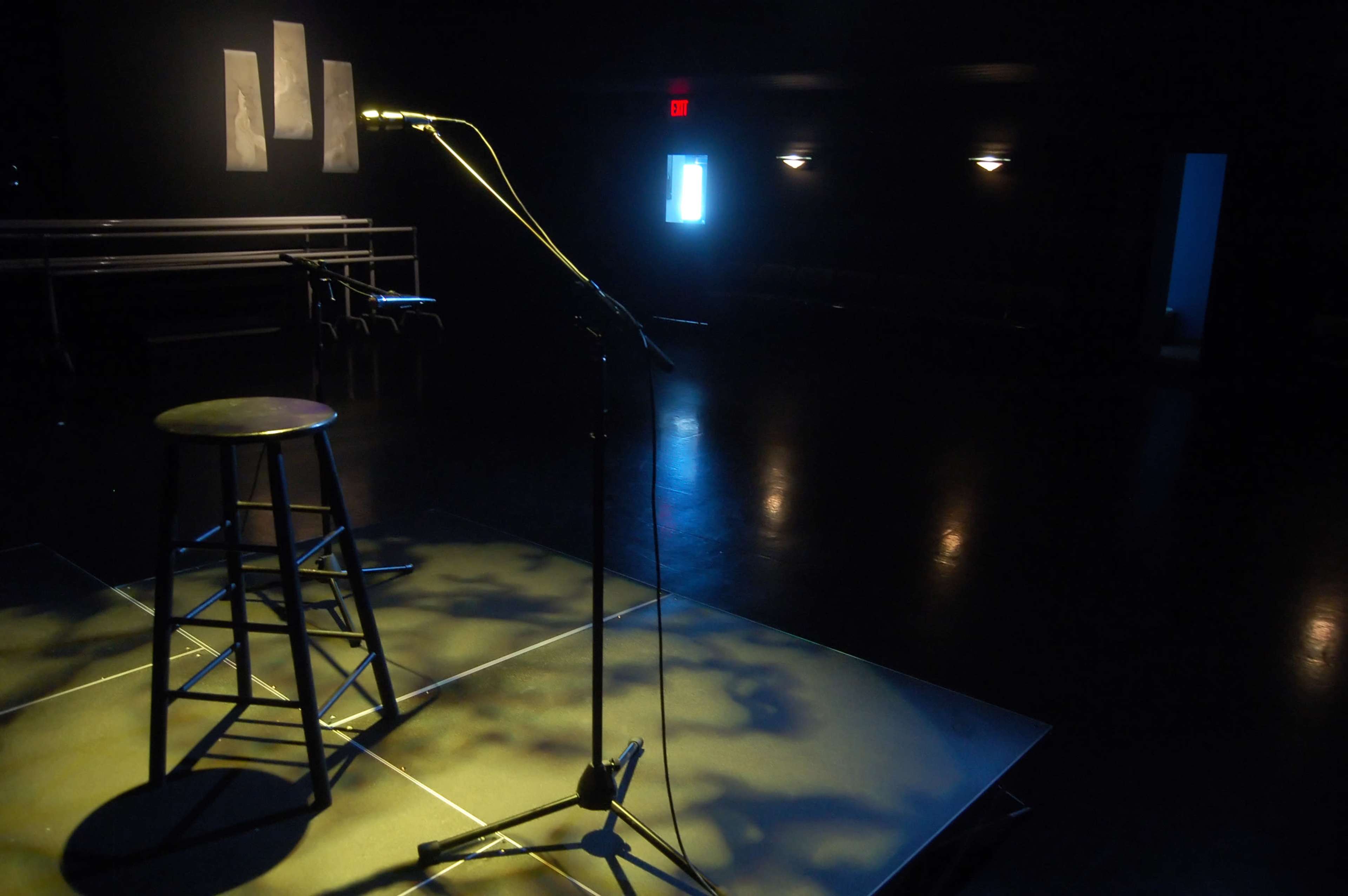 A spotlight illuminates an empty stage with a microphone stand and a stool, surrounded by a darkened room.