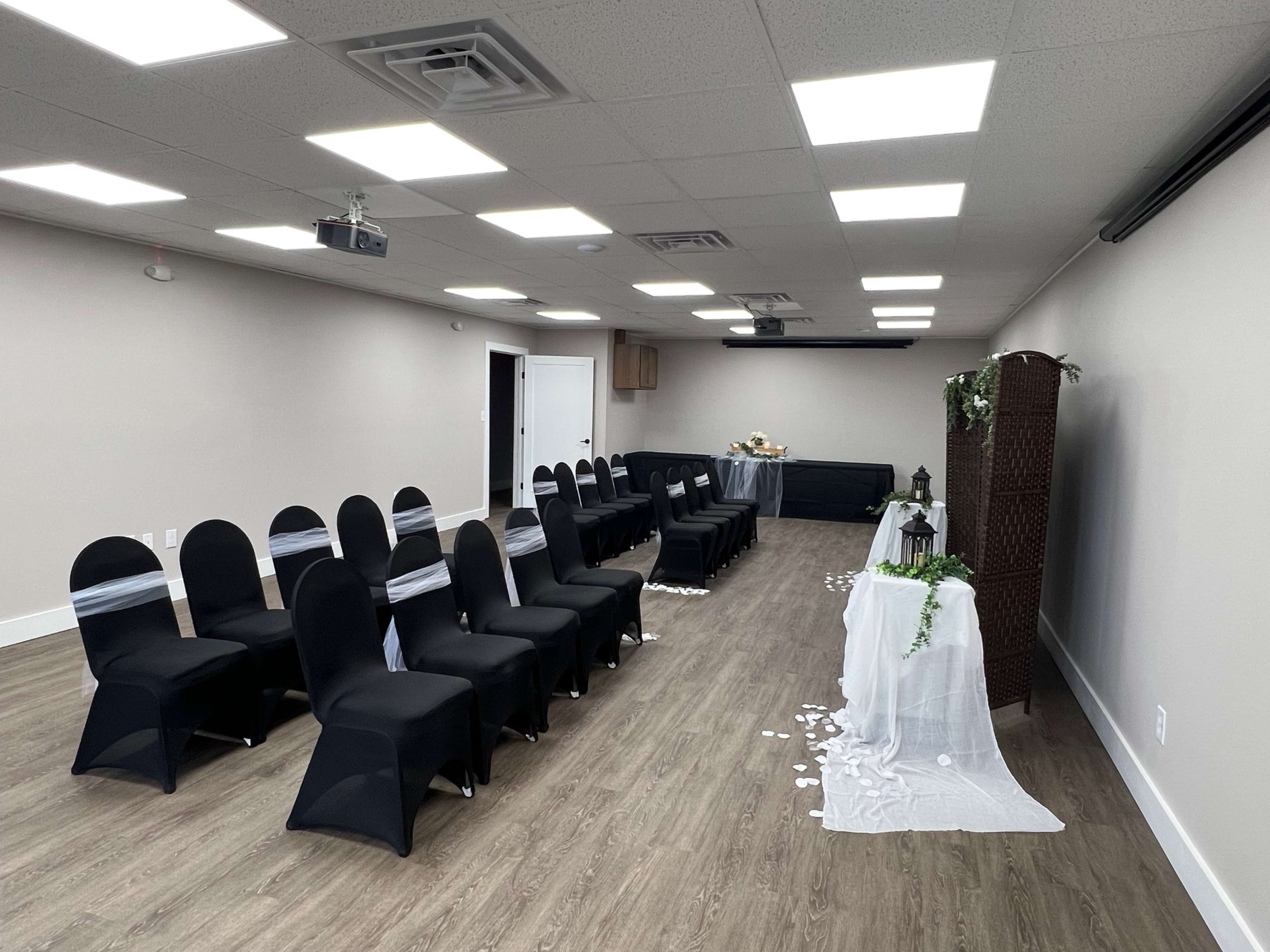 A wedding ceremony setup features rows of black chairs, a decorated table at the front, and scattered white petals on the floor.