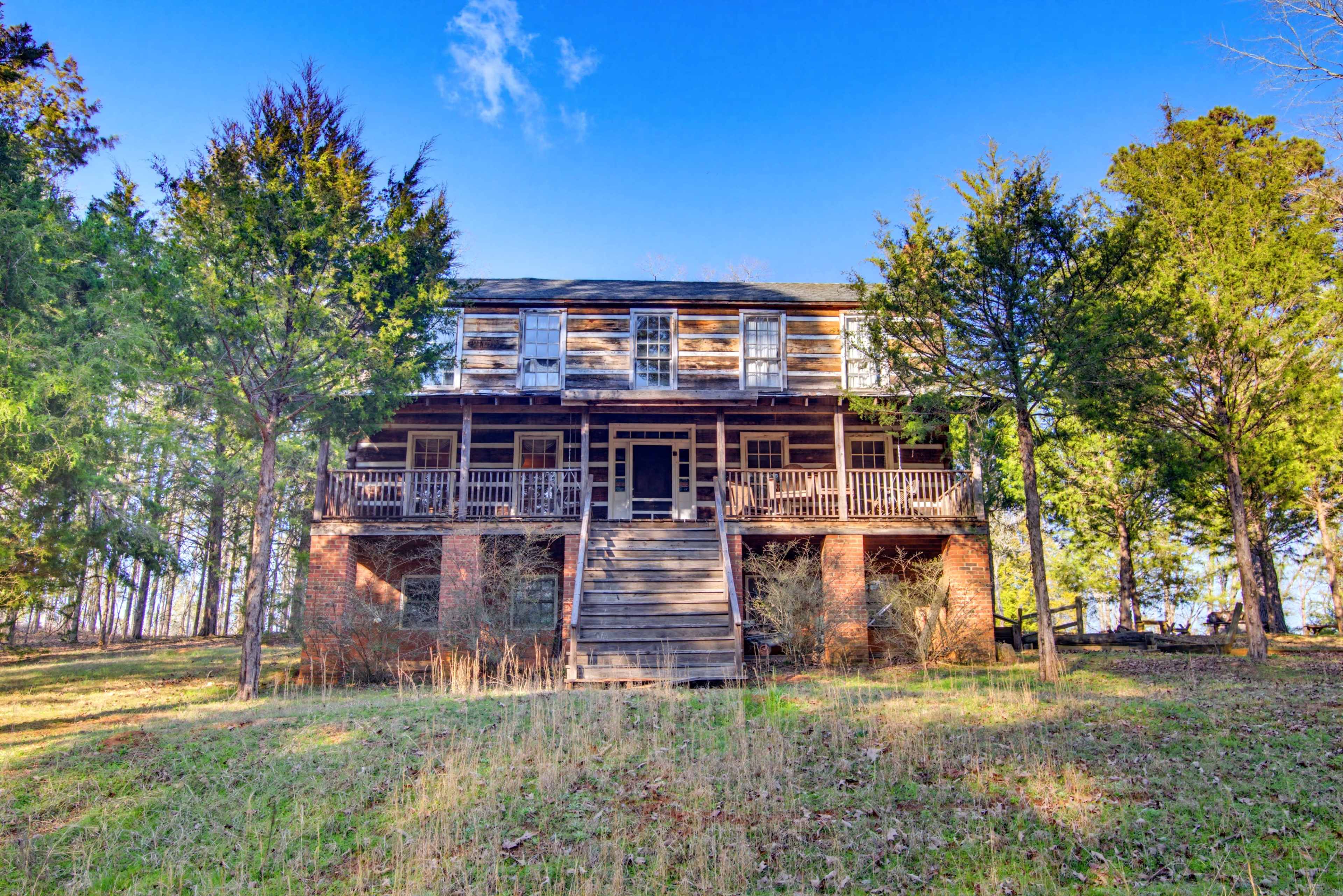 An old, two-story house with a porch and several windows is surrounded by trees in a grassy area.