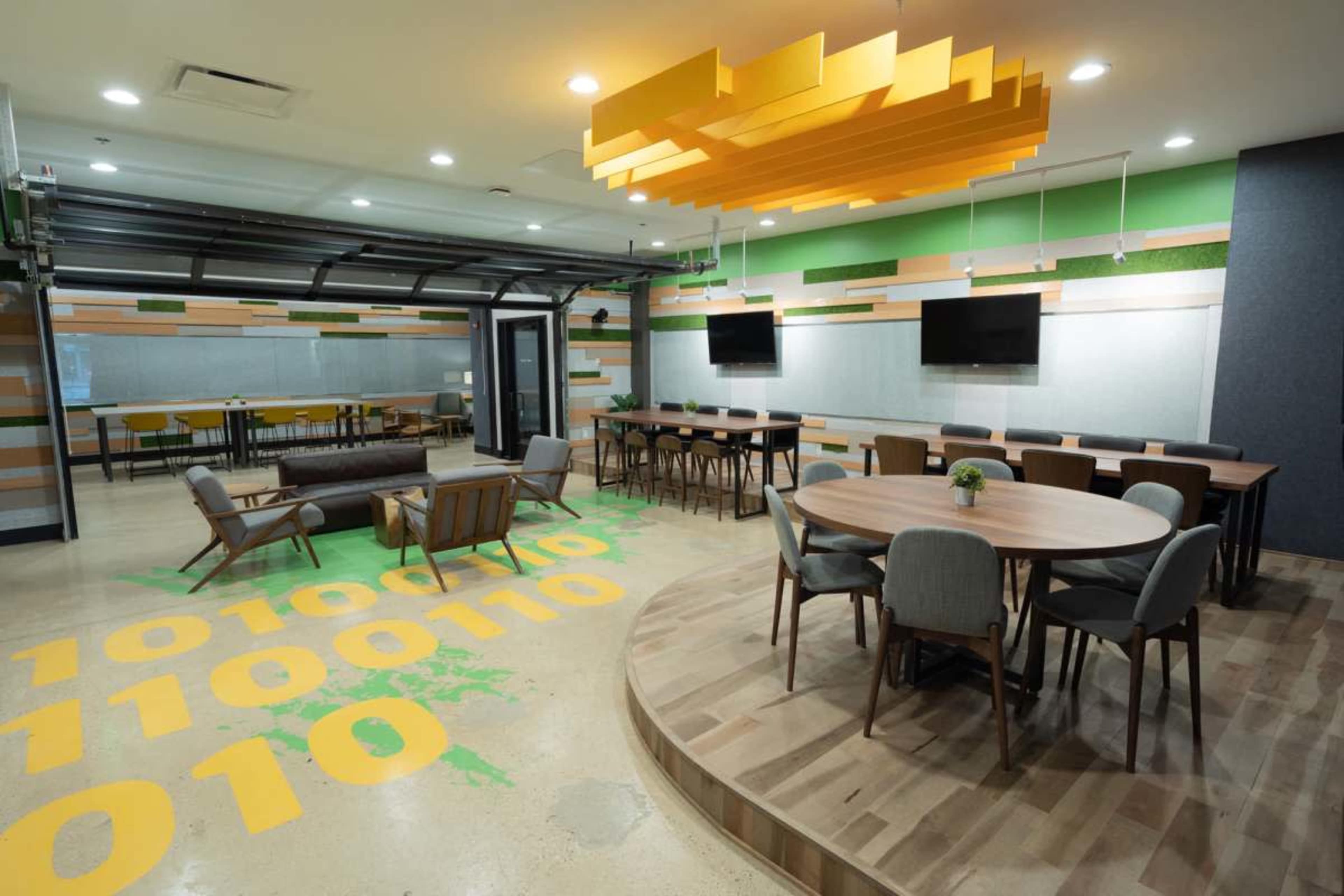 The image shows a modern, open office space featuring a large table surrounded by chairs, lounge seating, and a decorative ceiling installation, with binary numbers painted on the floor.