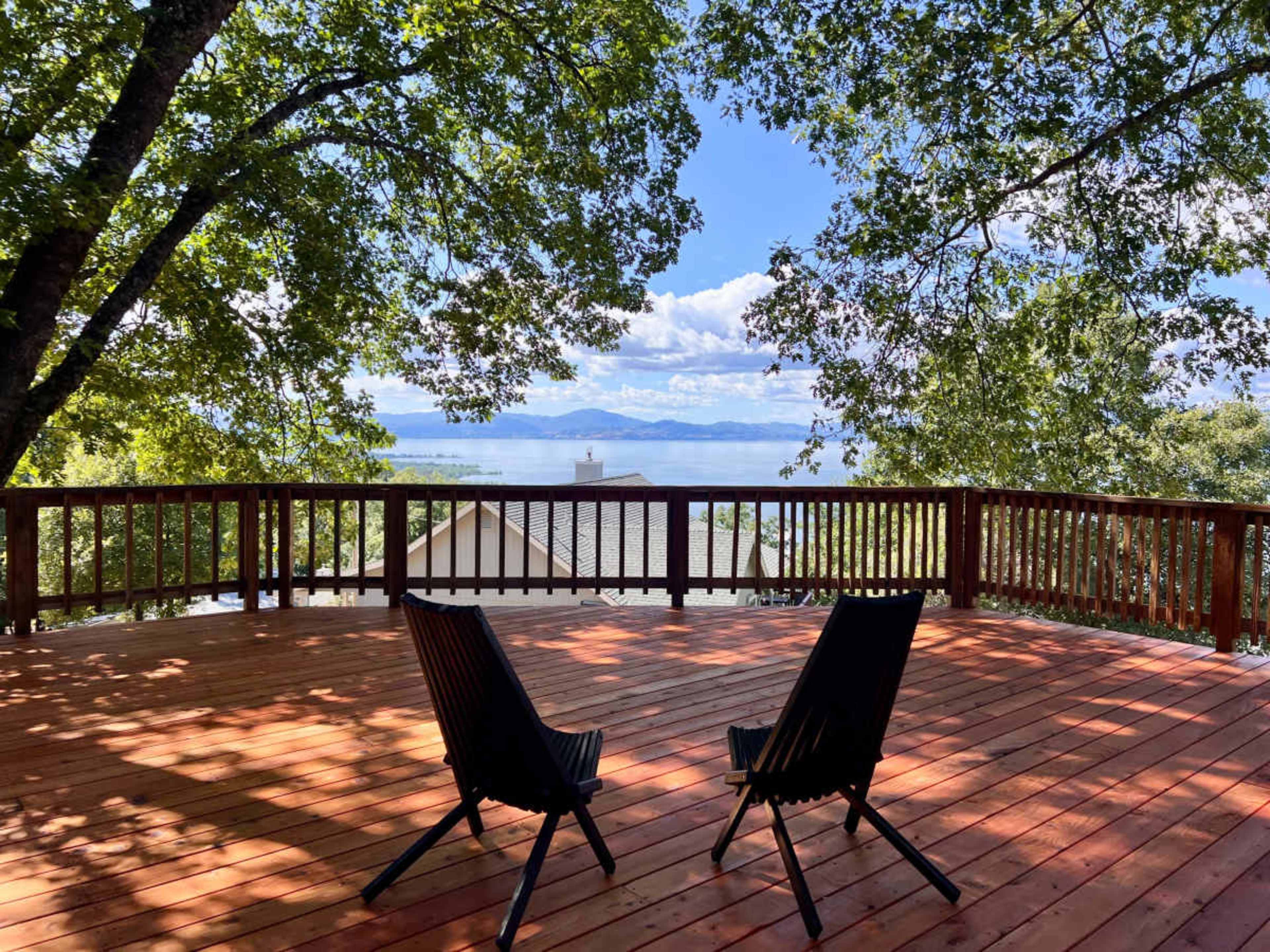 A wooden deck with two black chairs overlooks a lake surrounded by greenery under a blue sky with clouds.