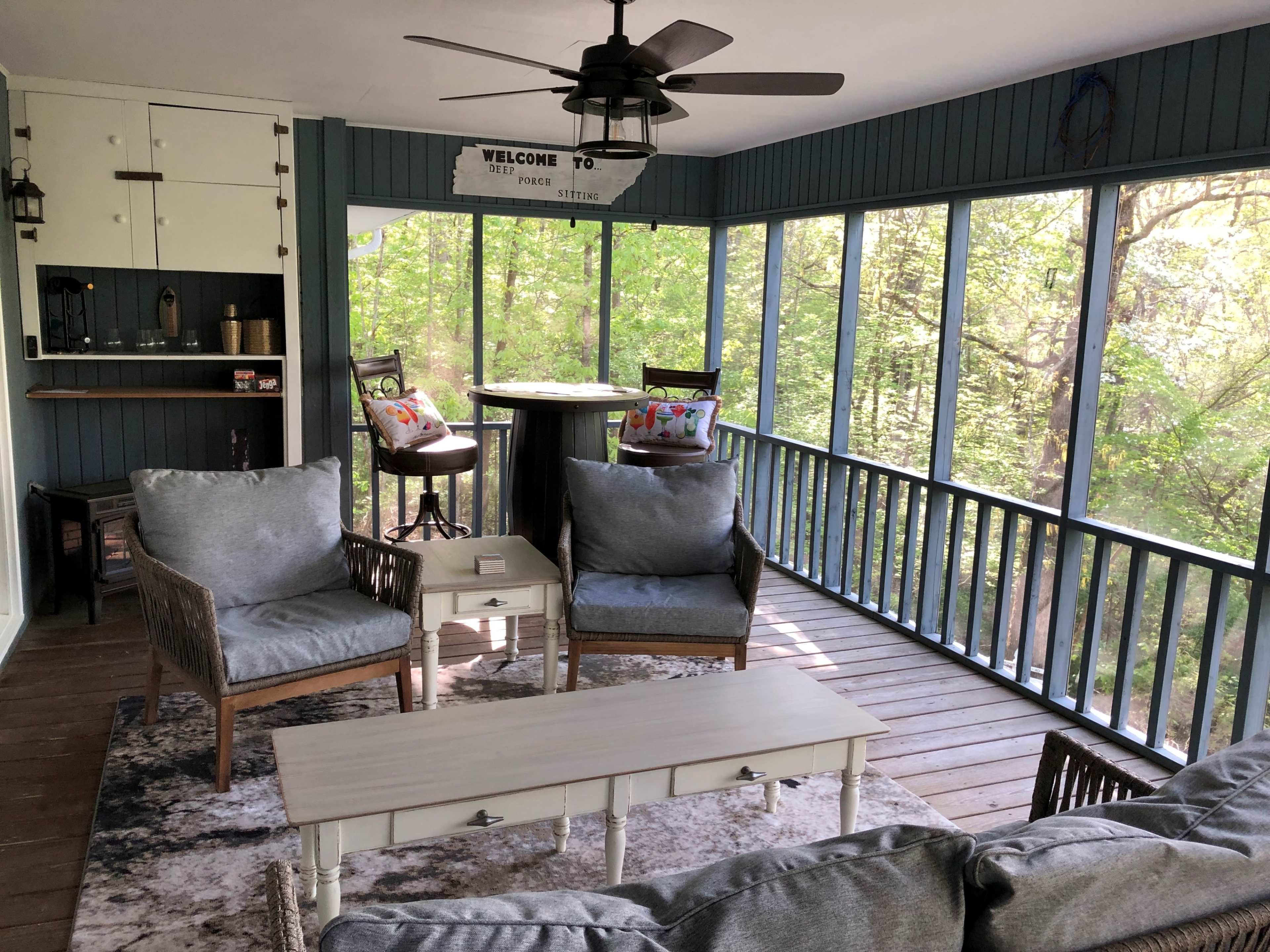 A screened-in porch featuring a seating area with grey cushioned chairs, a small table, a tall dining table with chairs, and a view of greenery outside.