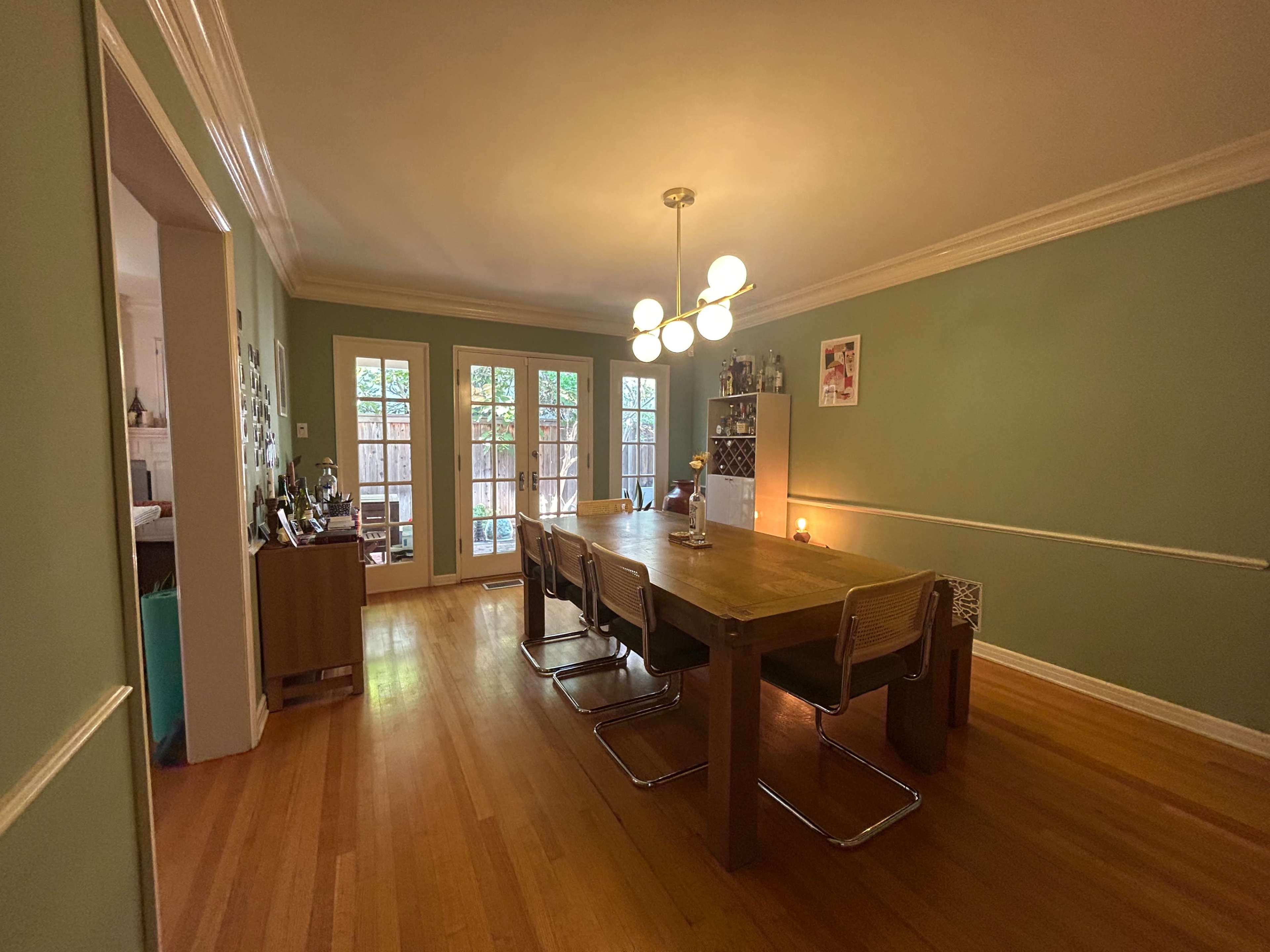 The image shows a spacious dining room with a wooden table and metal chairs, illuminated by a modern light fixture, and featuring doors that lead to an outdoor space.