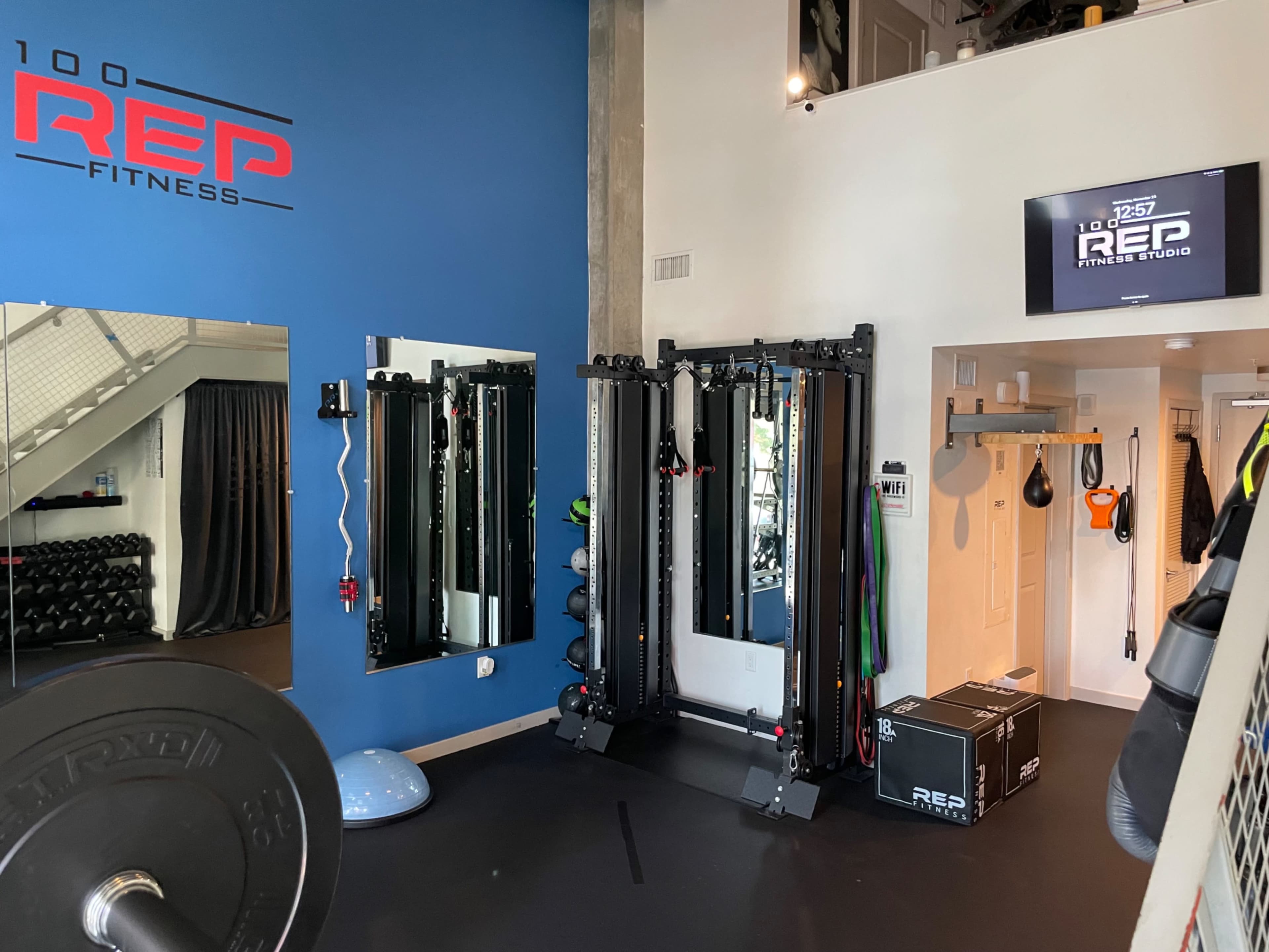 The image shows a fitness studio with exercise equipment, including weights, a cable machine, and a mirror, set against a blue wall featuring the logo of "100 REP FITNESS."
