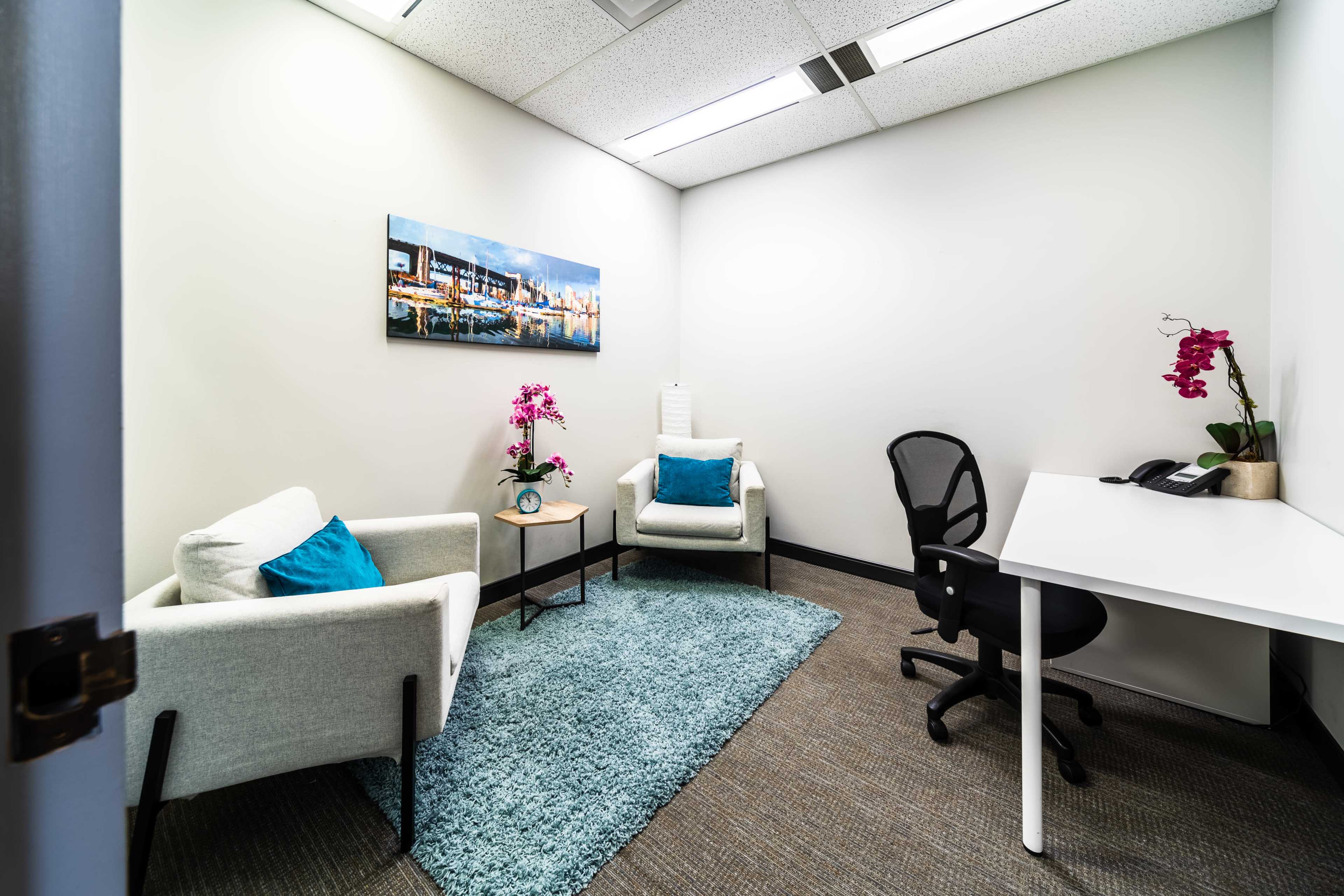 A small office space featuring two armchairs, a desk with a chair, a rug, and decorative plants, along with a painting on the wall.