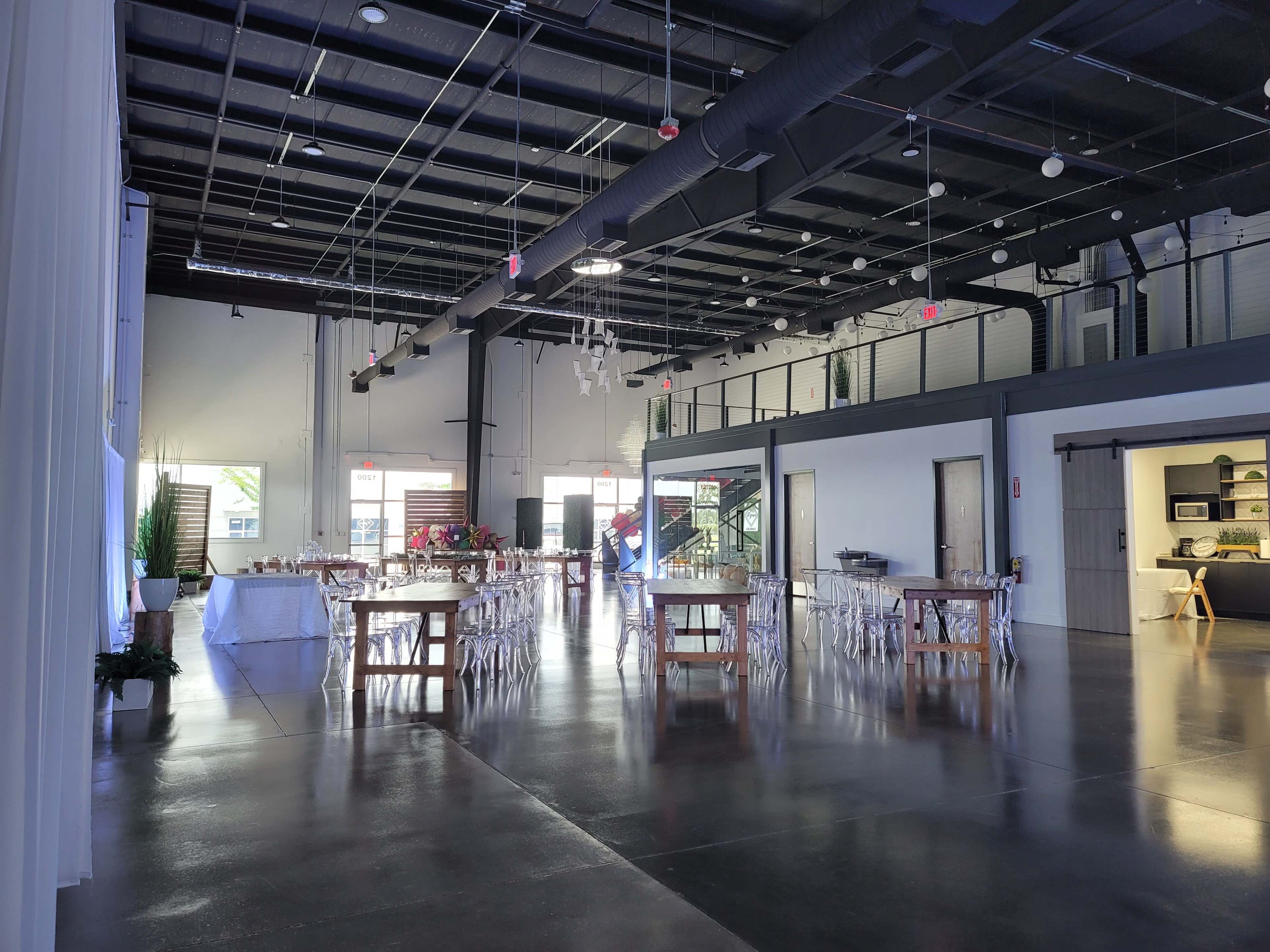 The image shows a spacious event venue with a polished concrete floor, several rectangular tables arranged for dining, and large windows allowing natural light to enter.