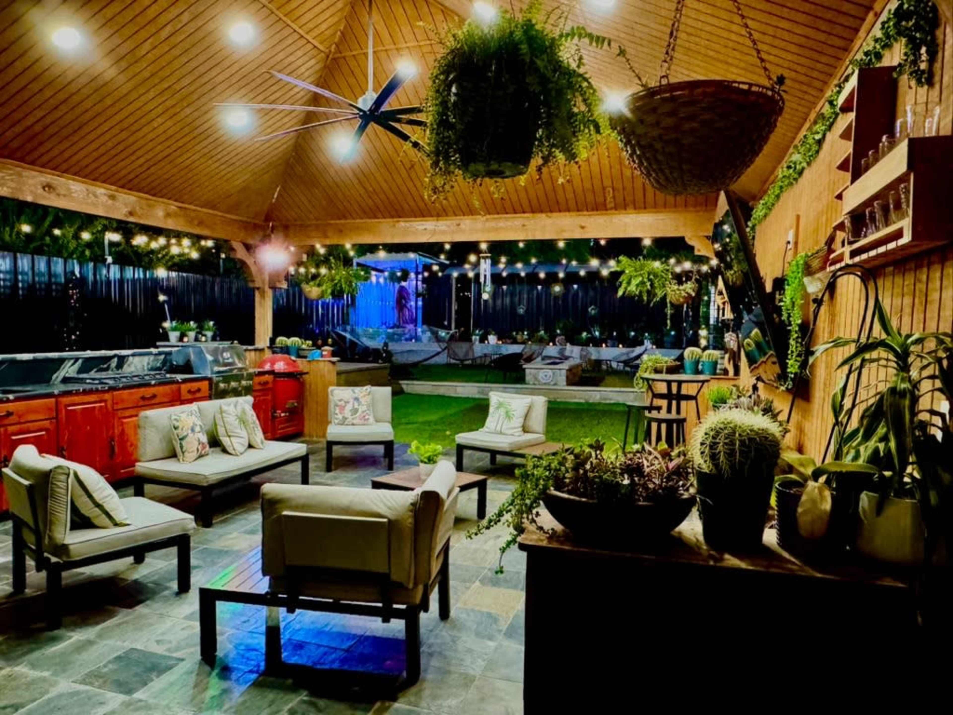 The image shows an outdoor patio area with a wooden ceiling, various seating arrangements, a kitchen space, and decorative plants hanging from the ceiling.