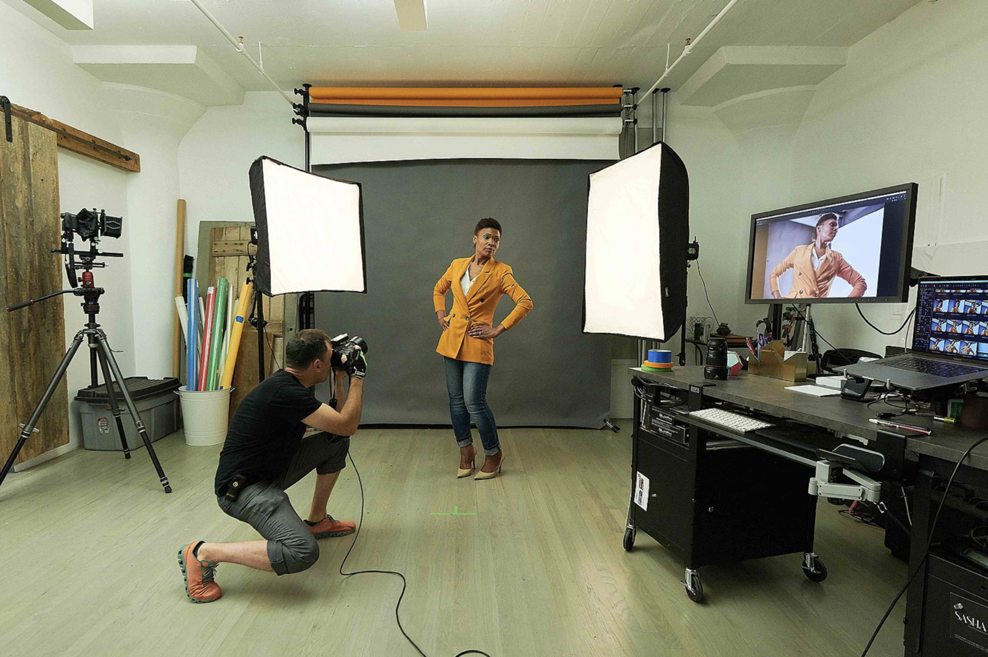 A photographer kneels to take a picture of a model posing in front of a backdrop in a photography studio.