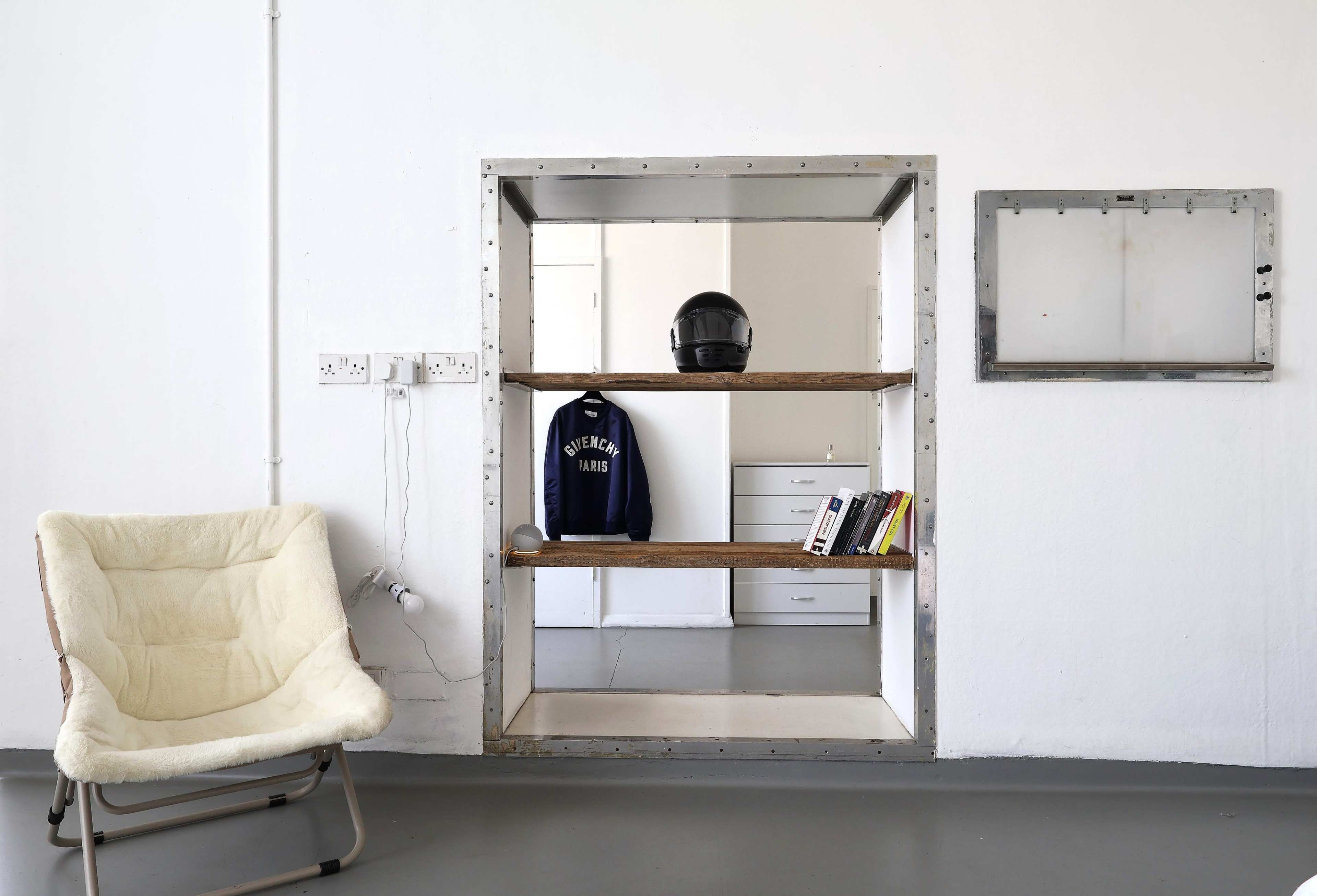 The image shows a minimalist room featuring an open wall shelf with a motorcycle helmet, a navy sweatshirt, and some books, alongside a cozy chair and kitchen area in the background.