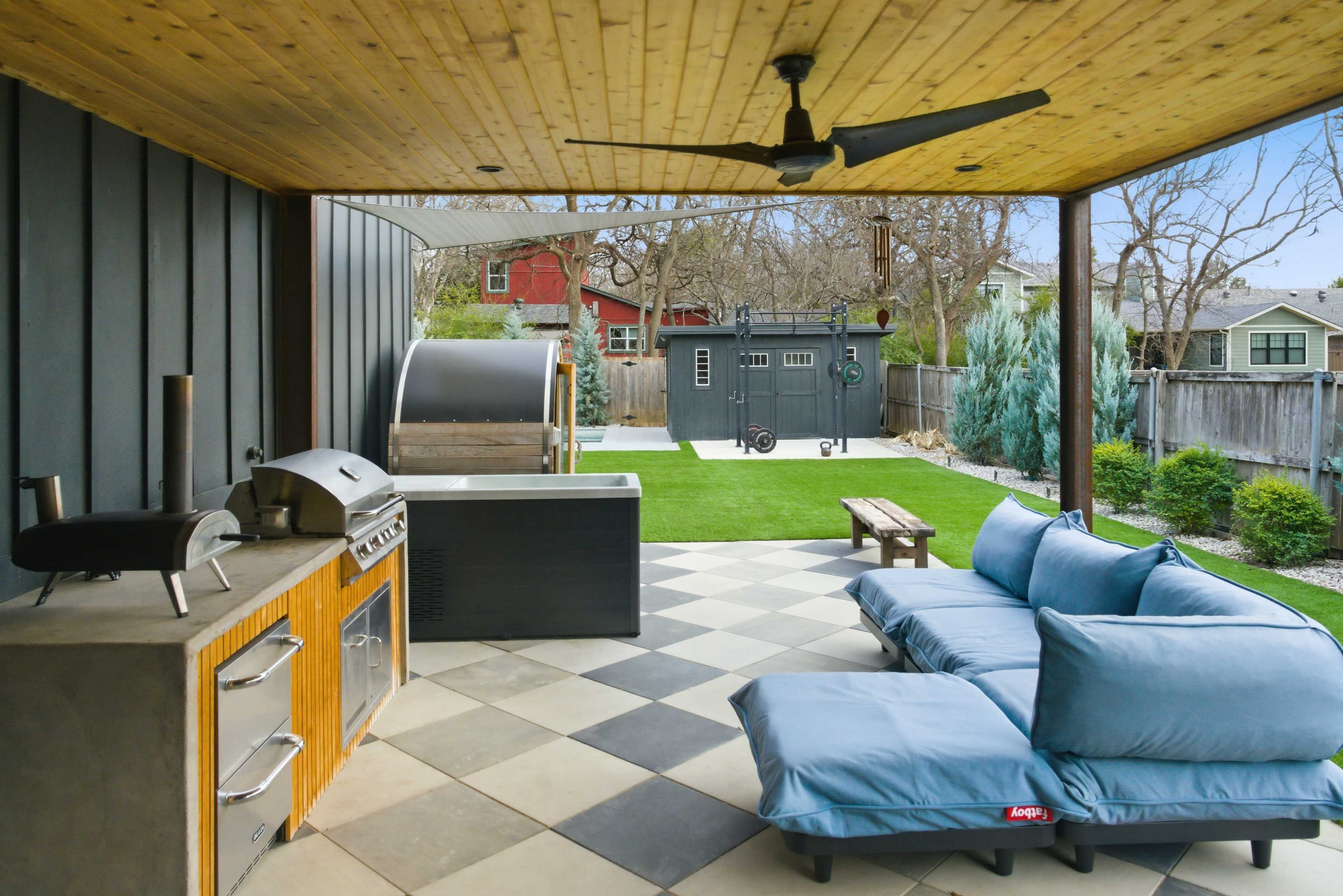 The image shows an outdoor patio area featuring a grill, a hot tub, and a couch set against a backdrop of a well-maintained lawn and garden structures.