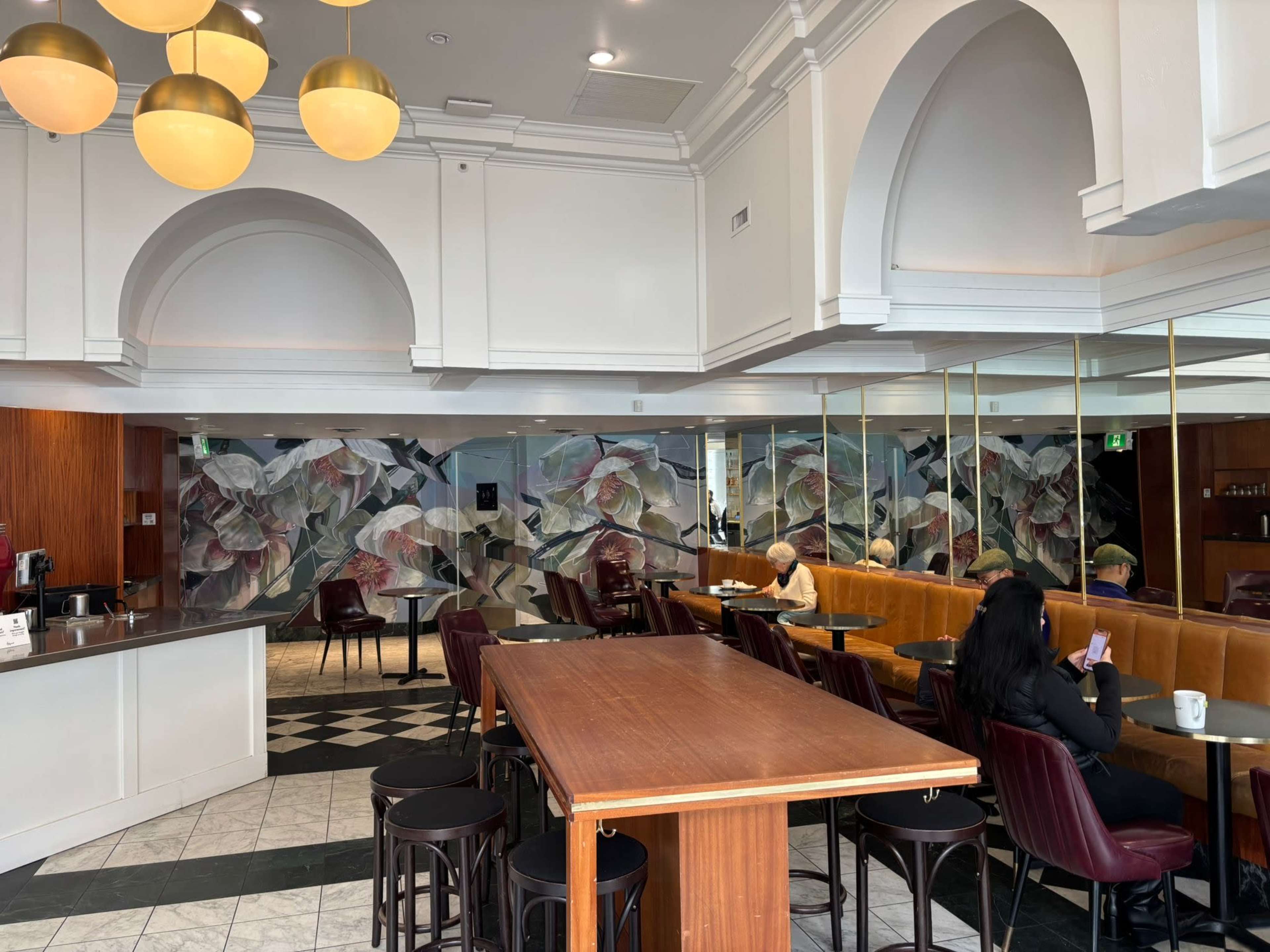The image shows a modern café interior featuring a wooden counter, dark chairs, and a large mural of flowers on the wall.