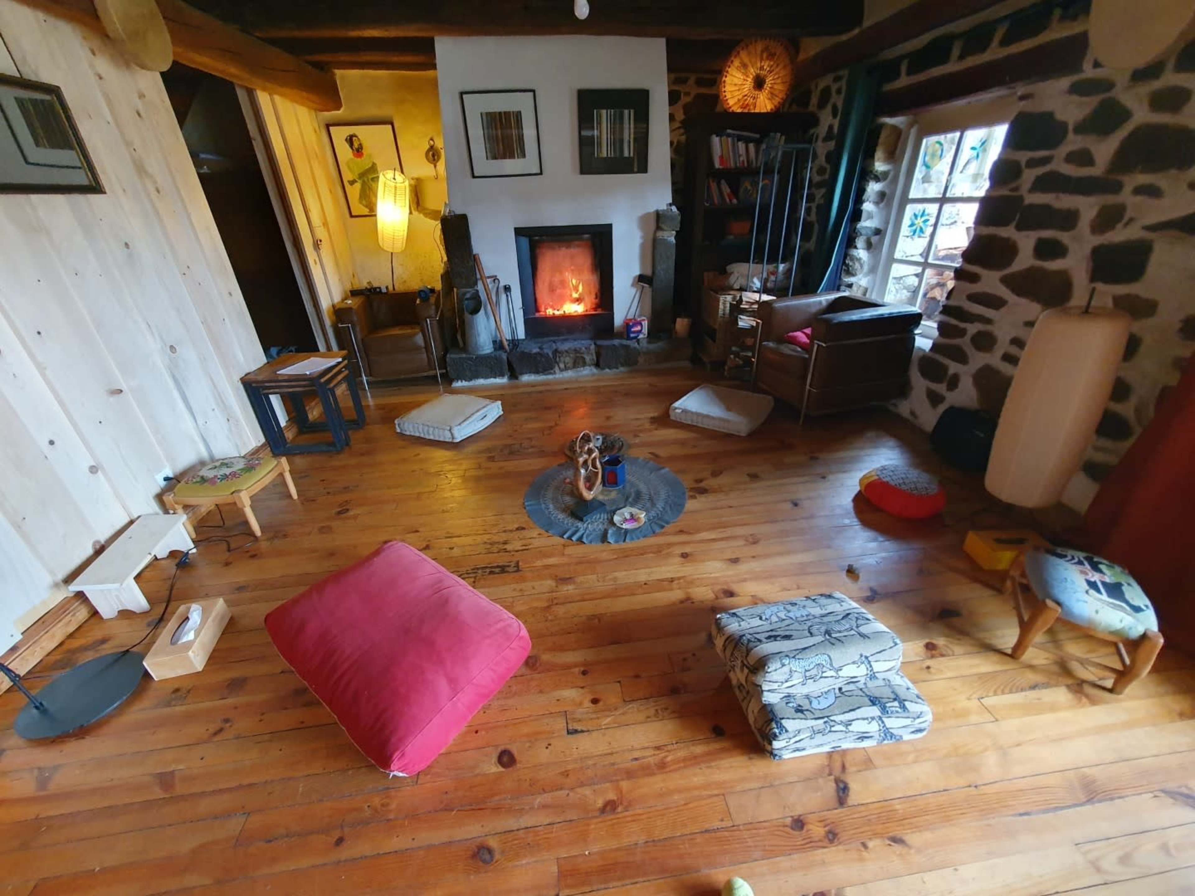 The image shows a cozy living room with wooden floors, a fireplace, assorted seating cushions, and bookshelves against the walls.