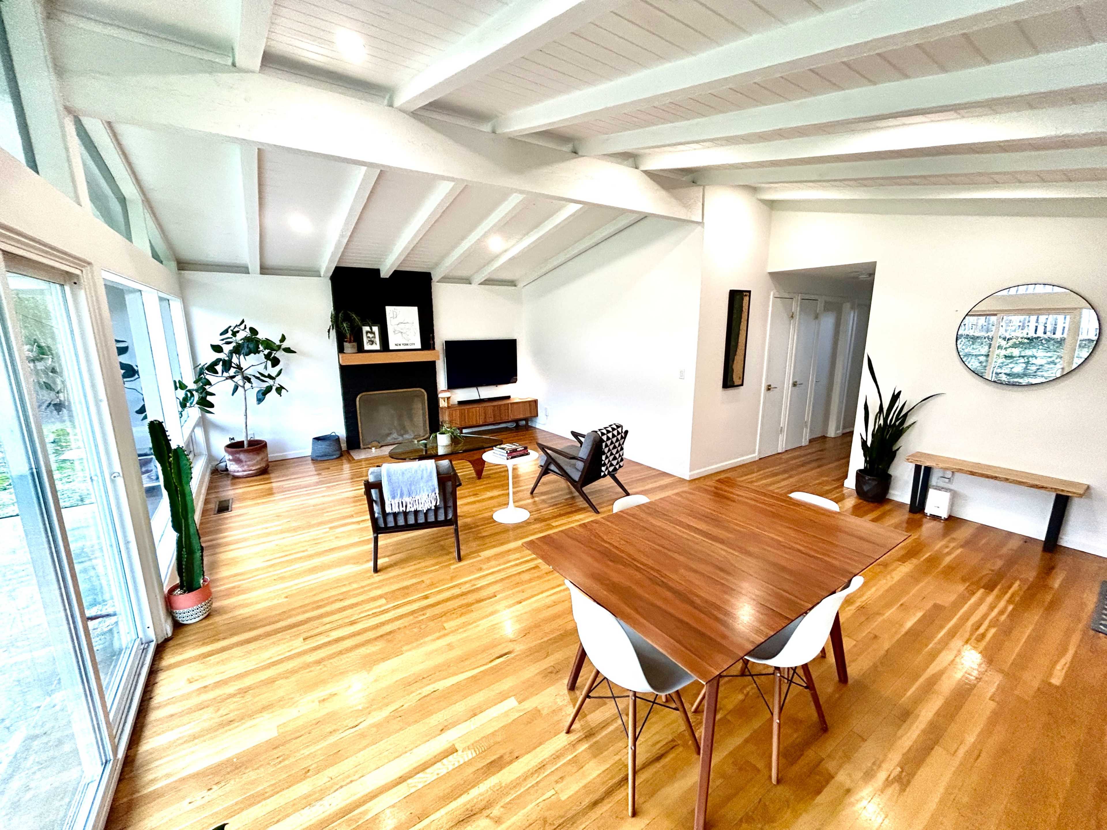 A spacious, well-lit living area featuring a wooden dining table, a fireplace, and large windows that offer a view of the outdoors.
