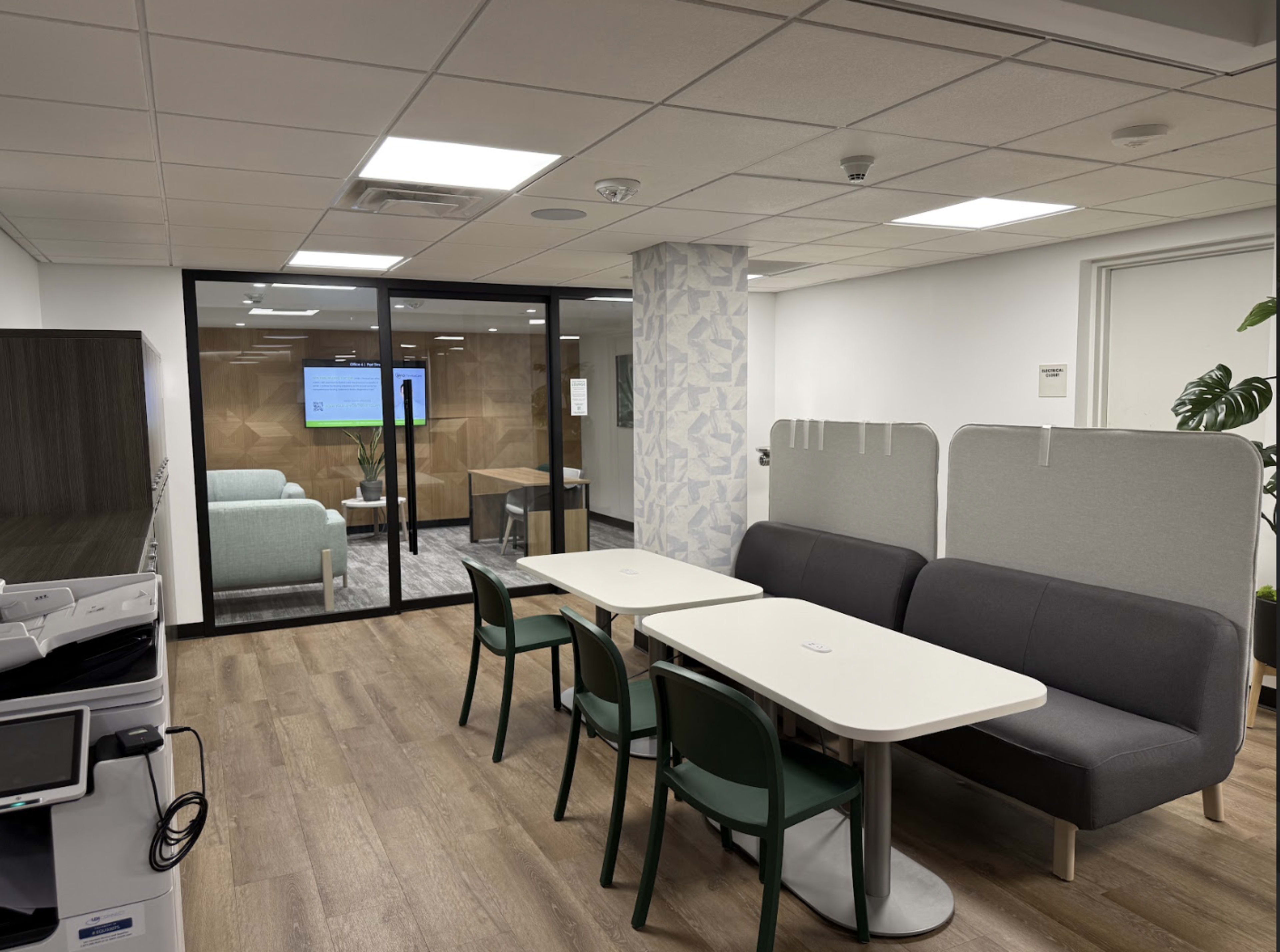 The image shows a modern office space featuring a seating area with a gray sofa, tables, and chairs, alongside a glass-walled meeting room in the background.