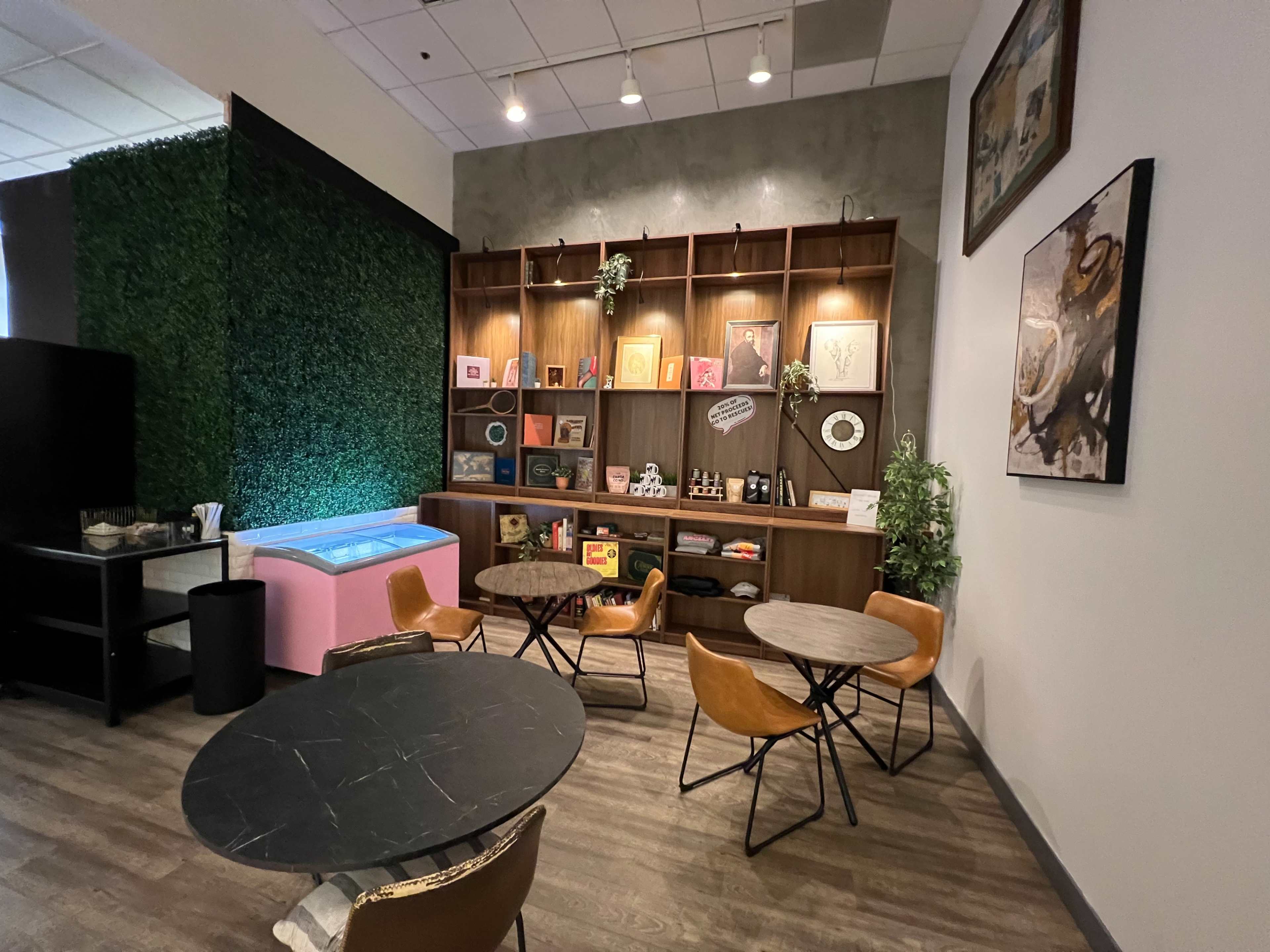 The image shows a cozy café area featuring two circular tables with brown chairs, a display shelf filled with colorful items, and a green wall on one side.