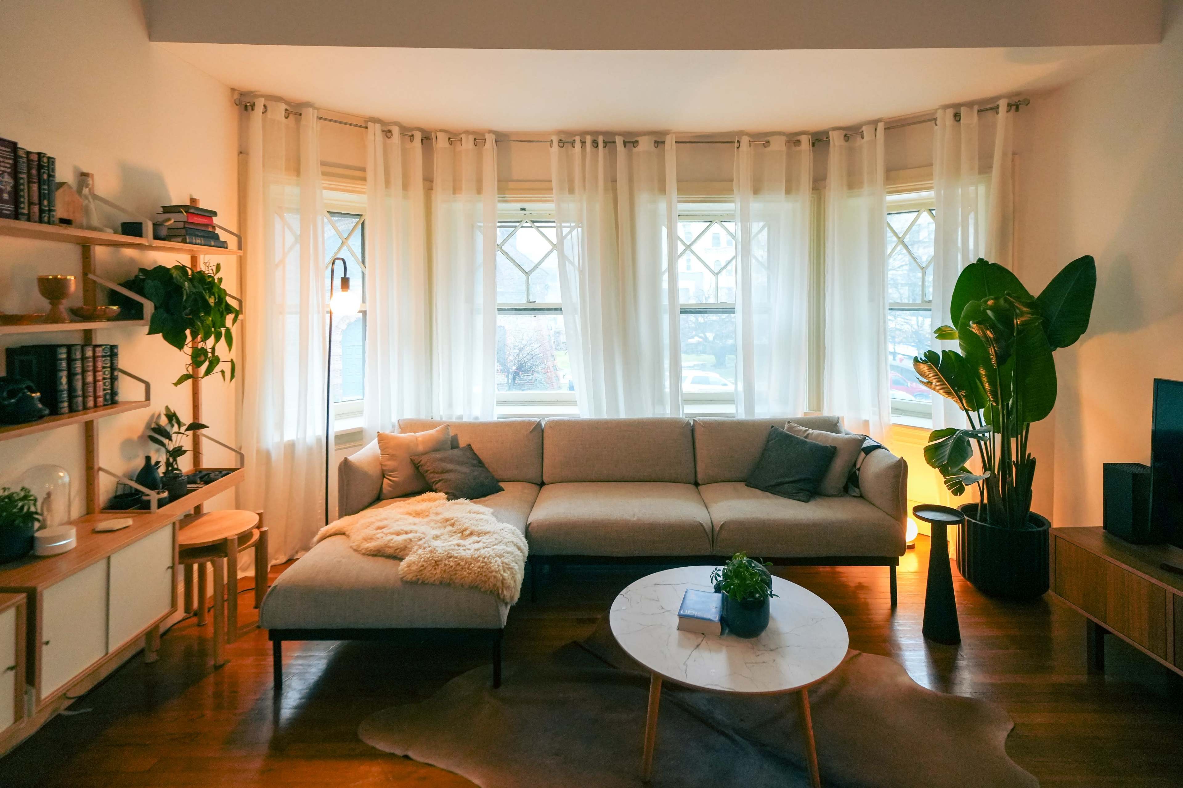 A cozy living room with a light-colored sectional sofa, large windows adorned with sheer curtains, a circular coffee table, and a potted plant.