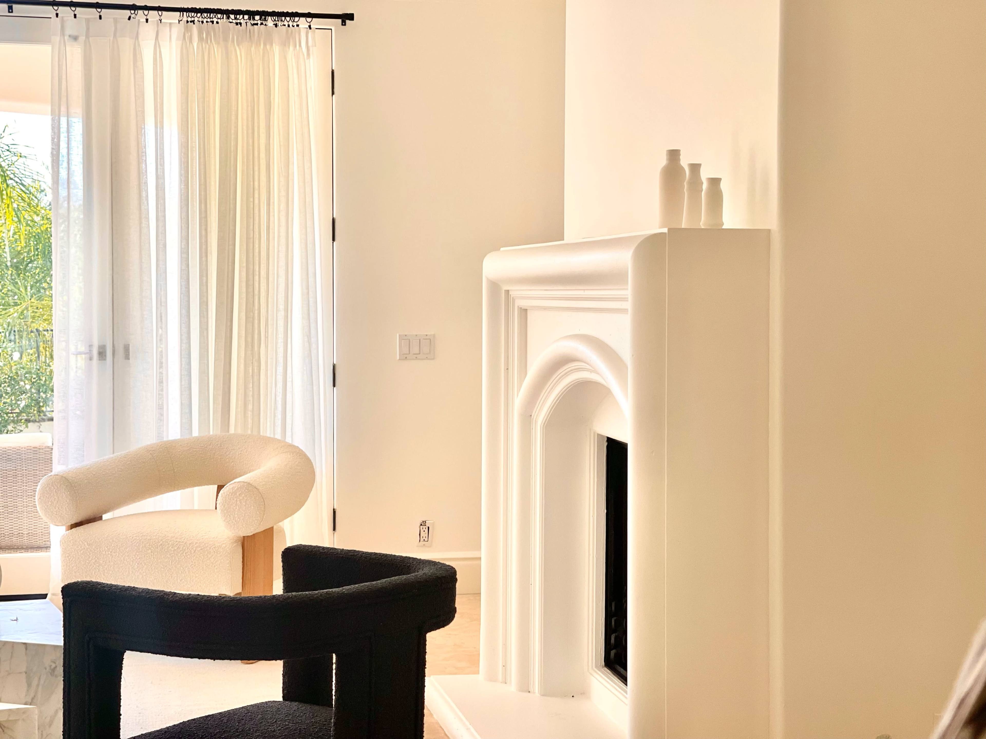A modern living room featuring a white fireplace, two distinct chairs, and sheer curtains allowing light to enter.