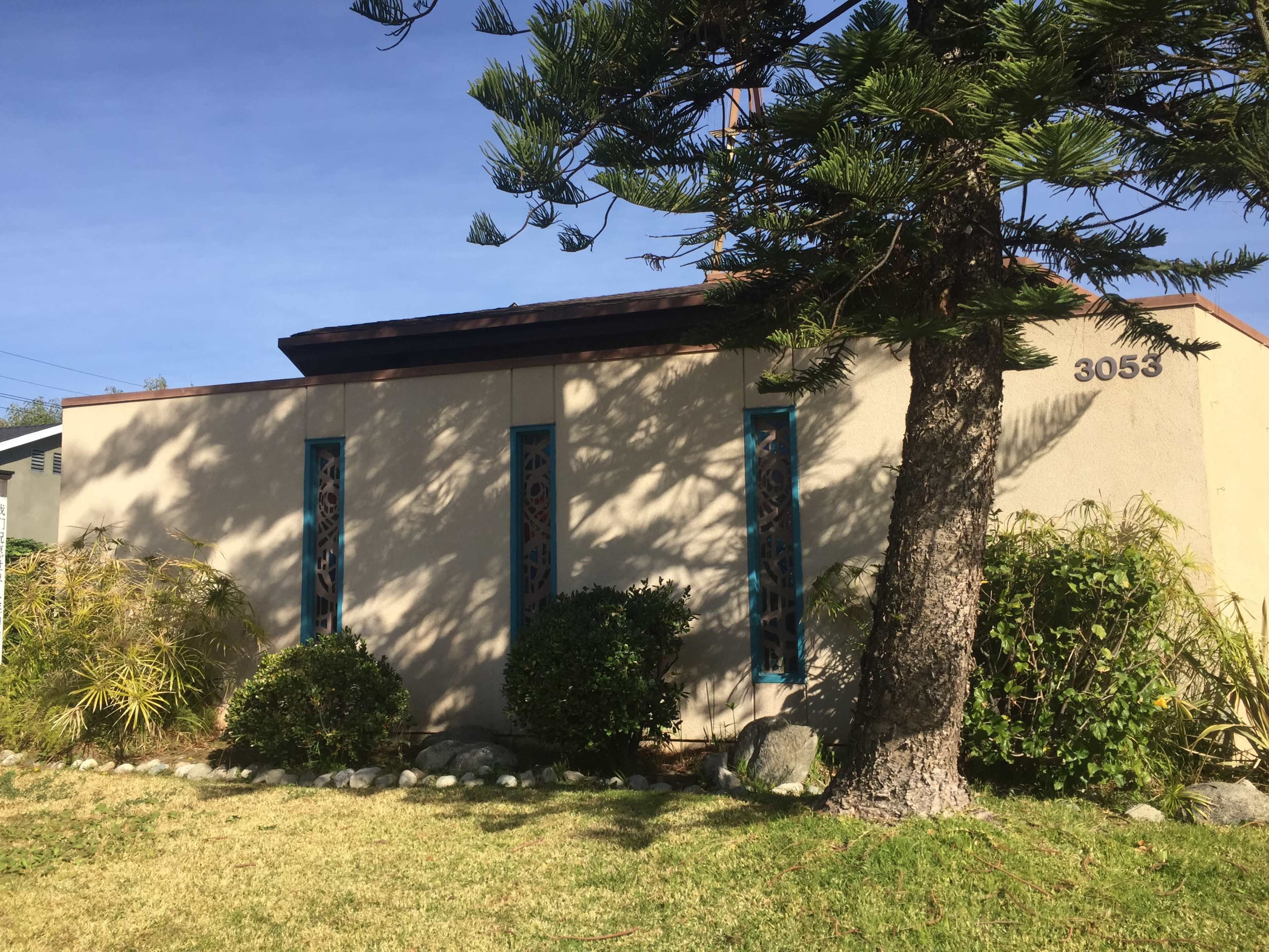 A single-story house with two blue-framed windows and a small yard containing green shrubs and a tree.