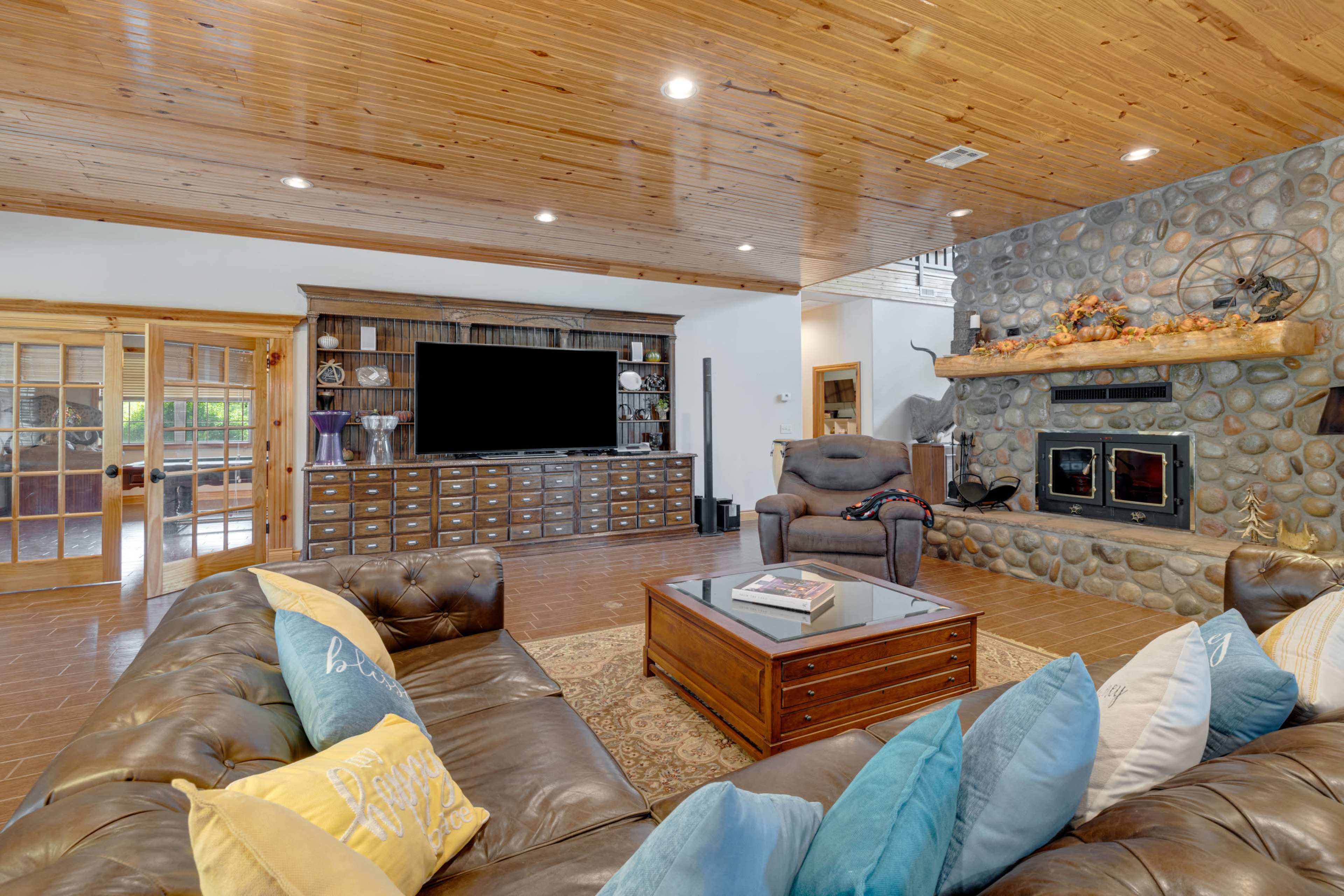 The spacious living room features a leather sectional sofa, a wooden coffee table, and a stone fireplace, with a large television mounted on the wall opposite built-in cabinetry.