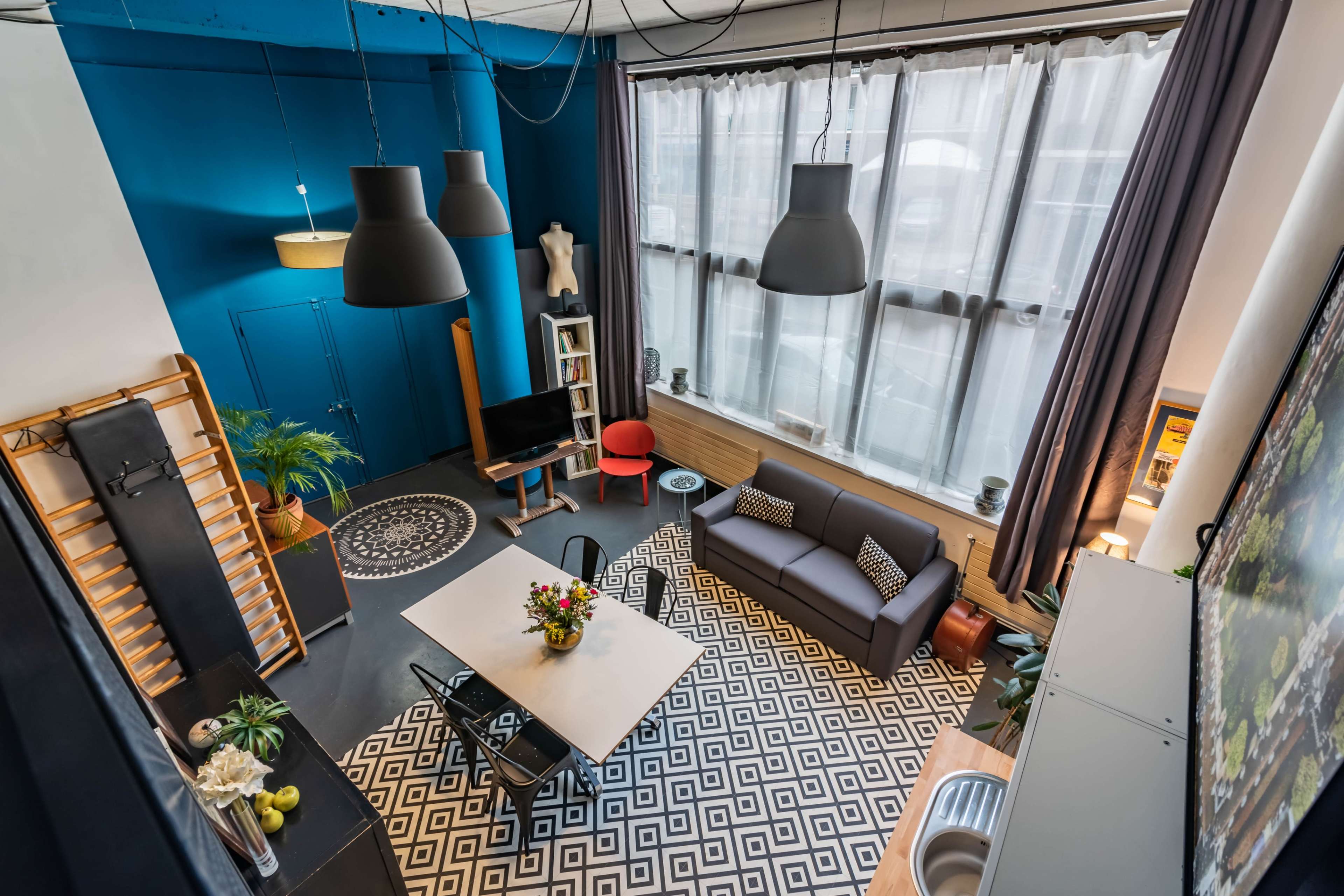 A modern loft interior features a large window, a patterned rug, a seating area with a sofa, a dining table, and various decorative elements.