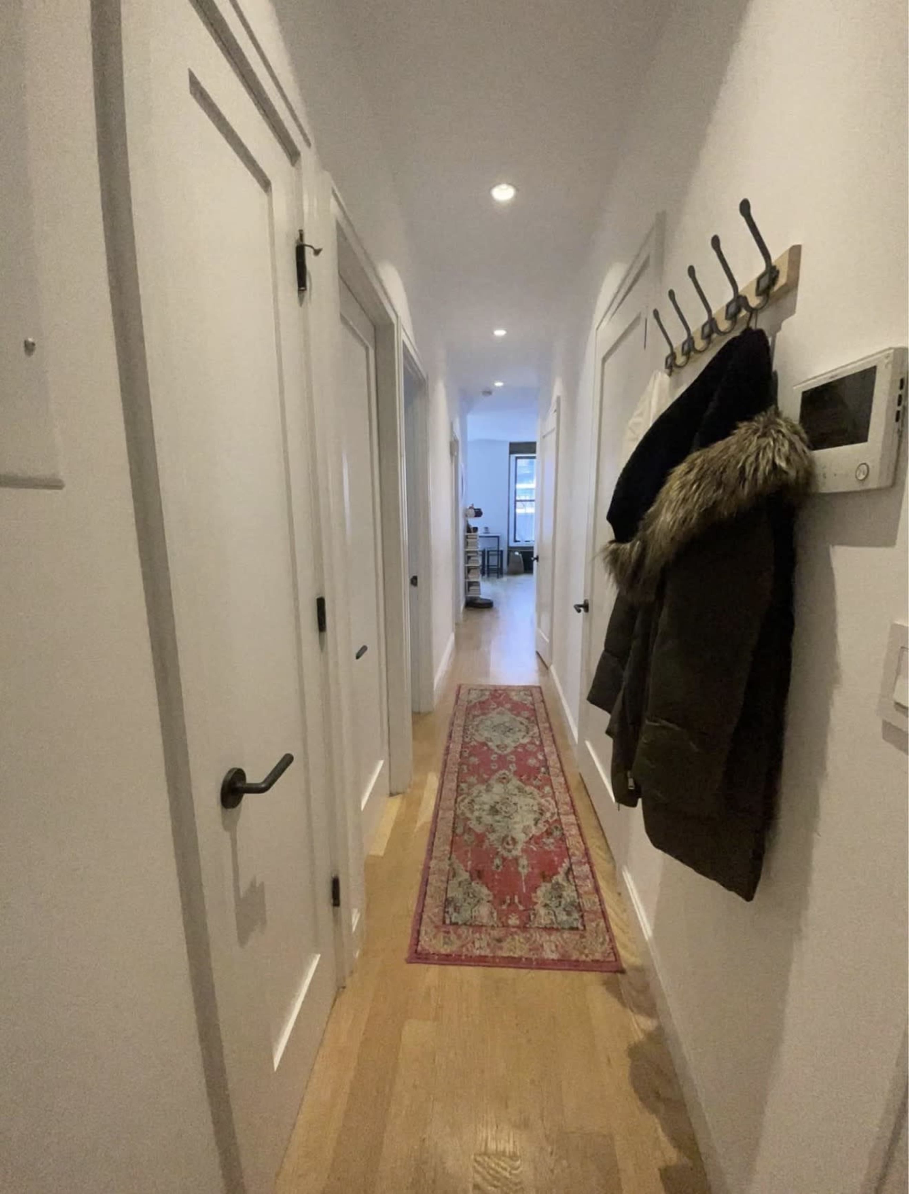 A narrow hallway features white walls, a patterned runner rug, hooks on the left for coats, and doors leading to rooms on either side.