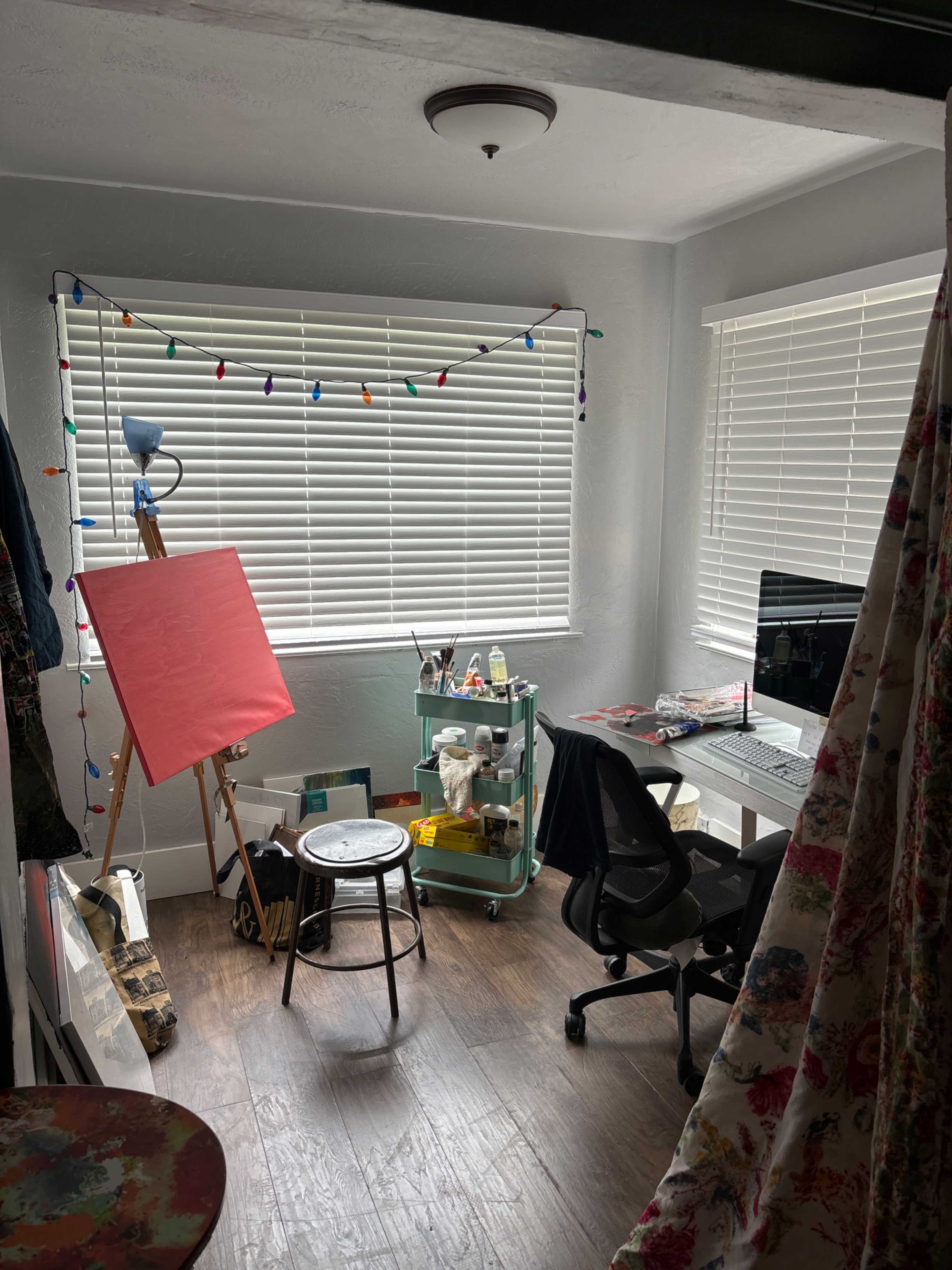 An artist's workspace featuring an easel with a red canvas, a computer desk, a rolling cart with art supplies, and string lights along the window.