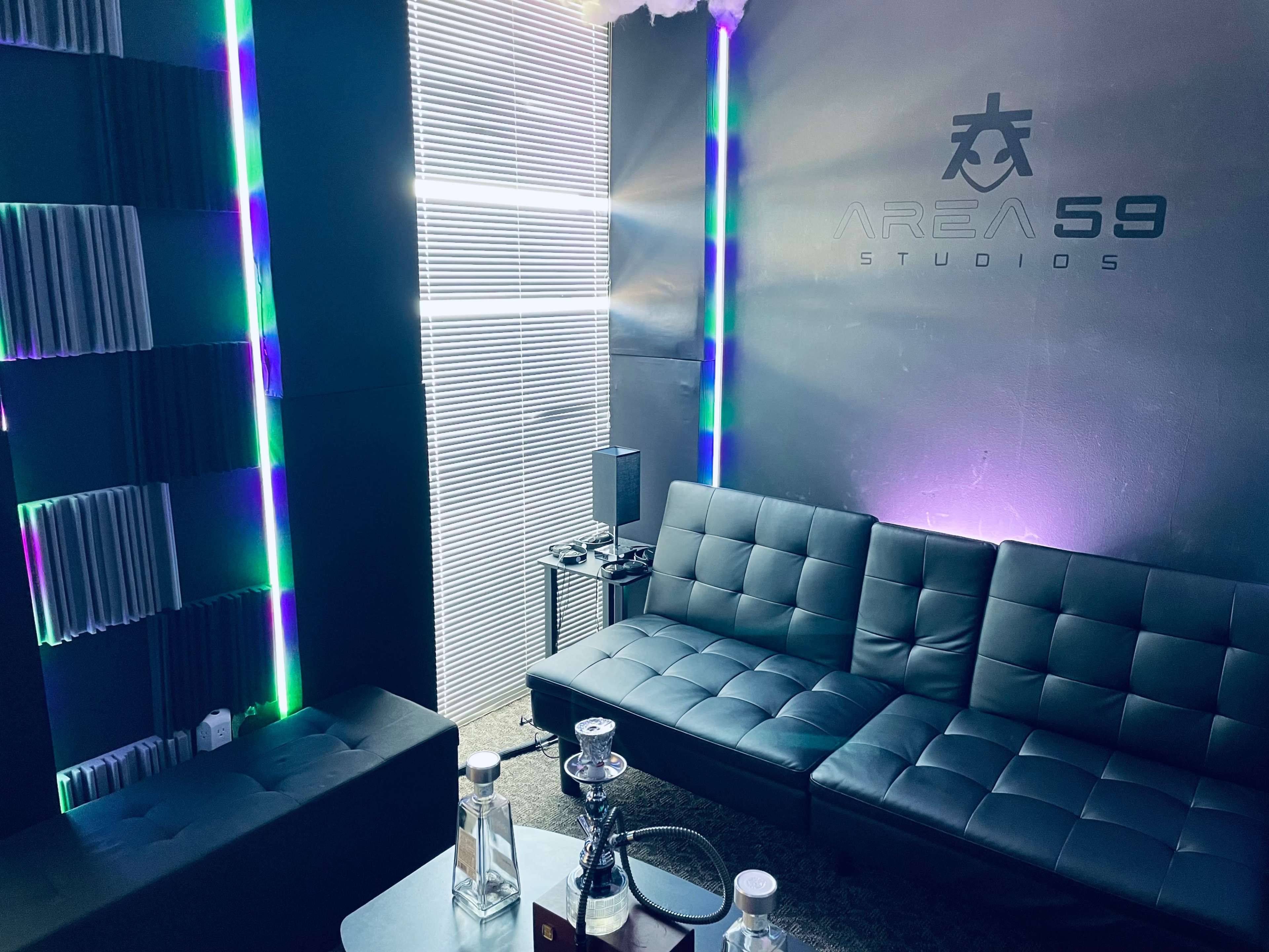 A modern studio lounge area with a black sectional sofa, ambient lighting, acoustic paneling on the walls, and branding for "Area 59 Studios."