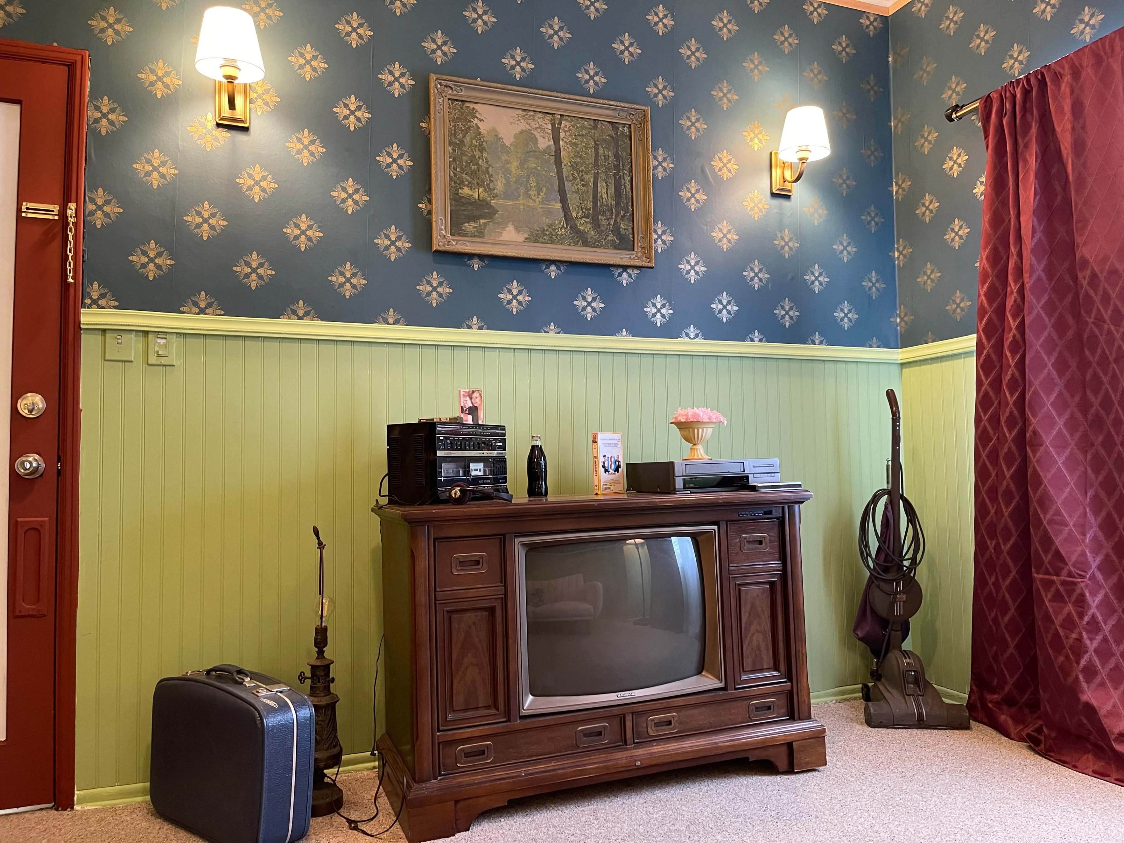 The scene features a retro living room with a wood cabinet housing a television, a small stereo system, a suitcase, and a vacuum cleaner, all set against a patterned blue and green wall and accented by wall sconces.