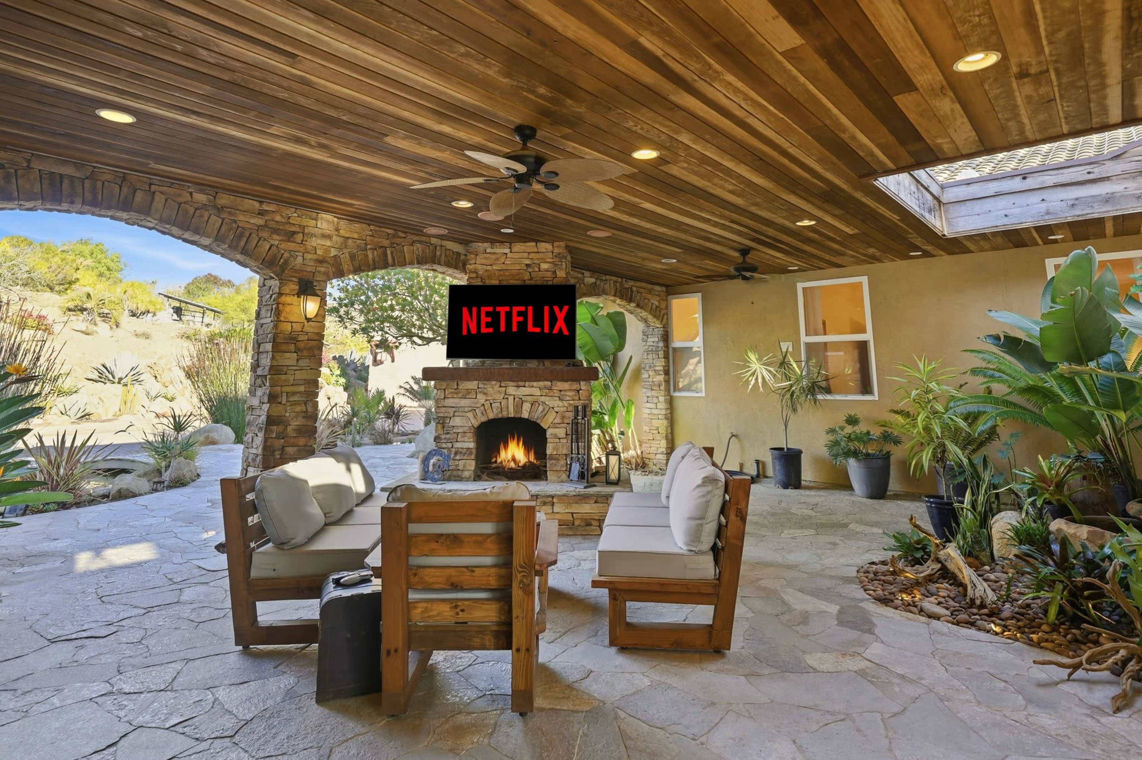 A stone patio features a fireplace surrounded by seating and tropical plants, beneath a wooden ceiling.