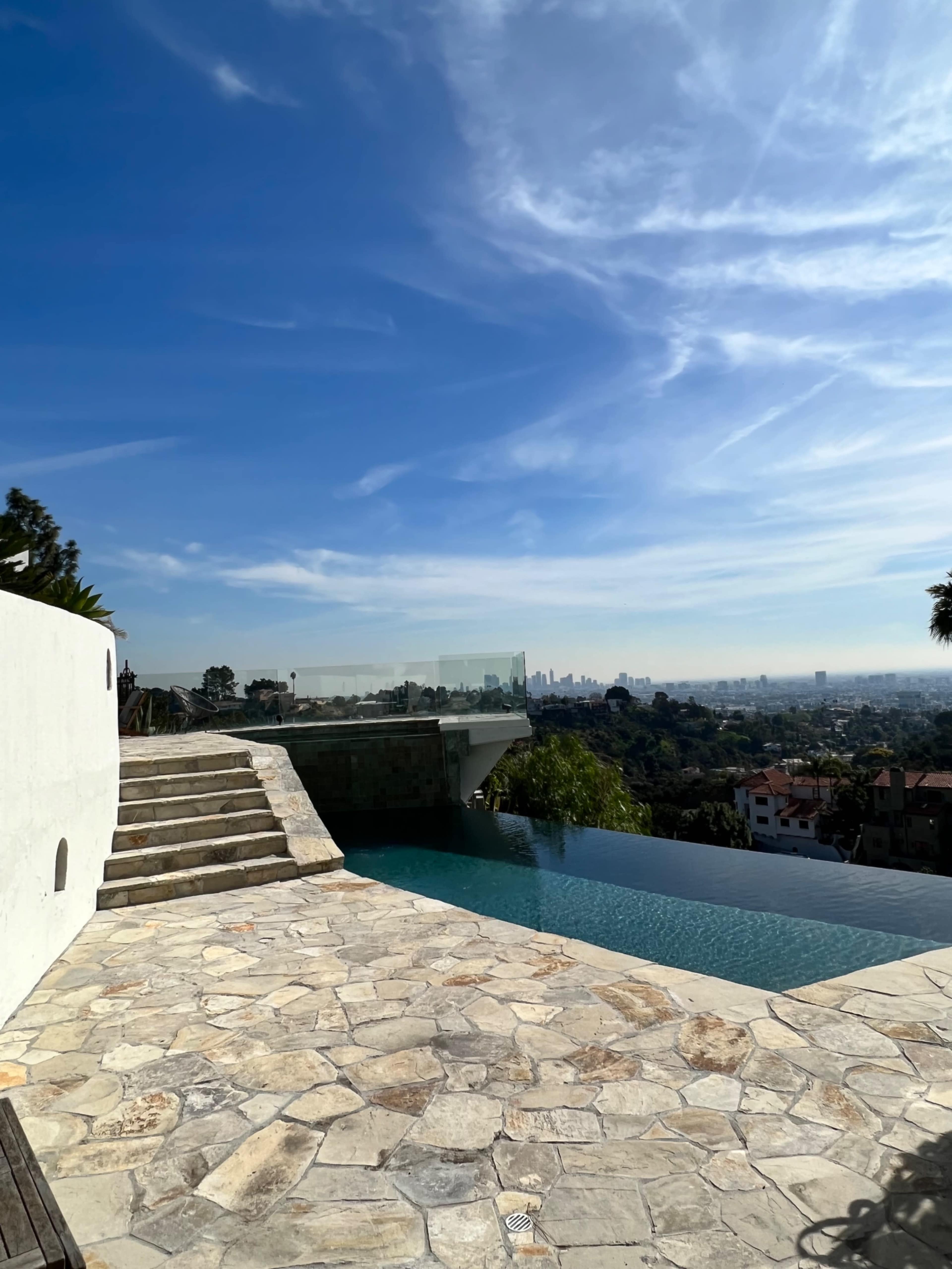 The image shows a modern infinity pool overlooking a city skyline under a clear blue sky.